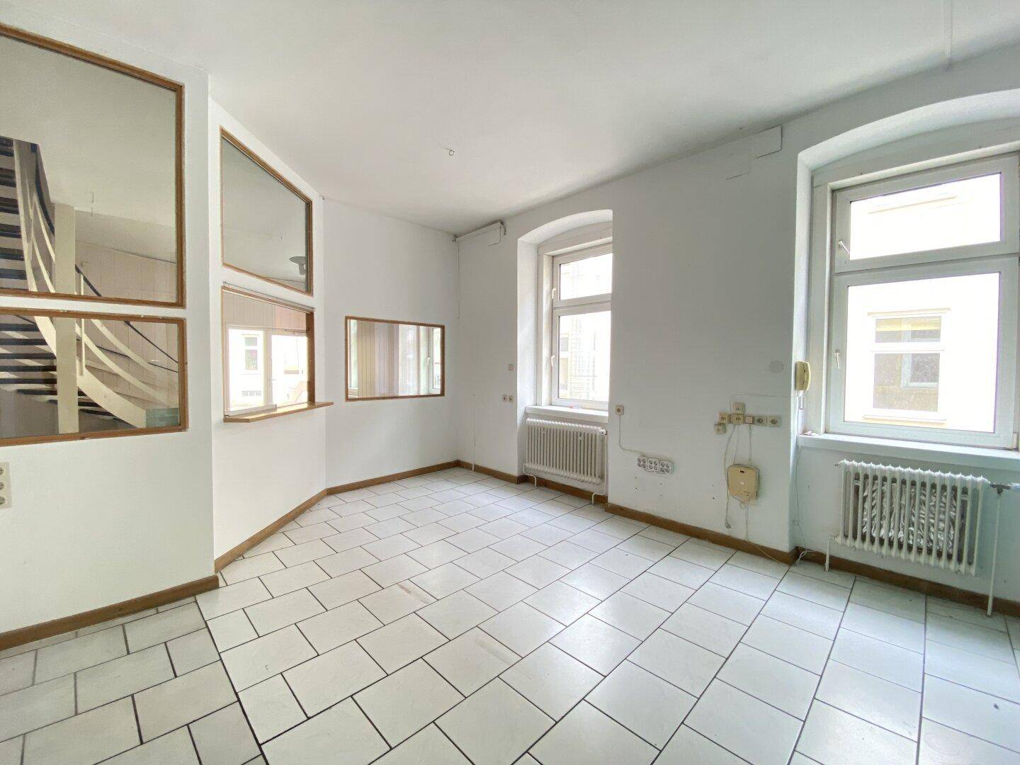 Heller Büroraum mit weißem Fliesenboden, zwei großen Fenstern und internen Glasabtrennungen.