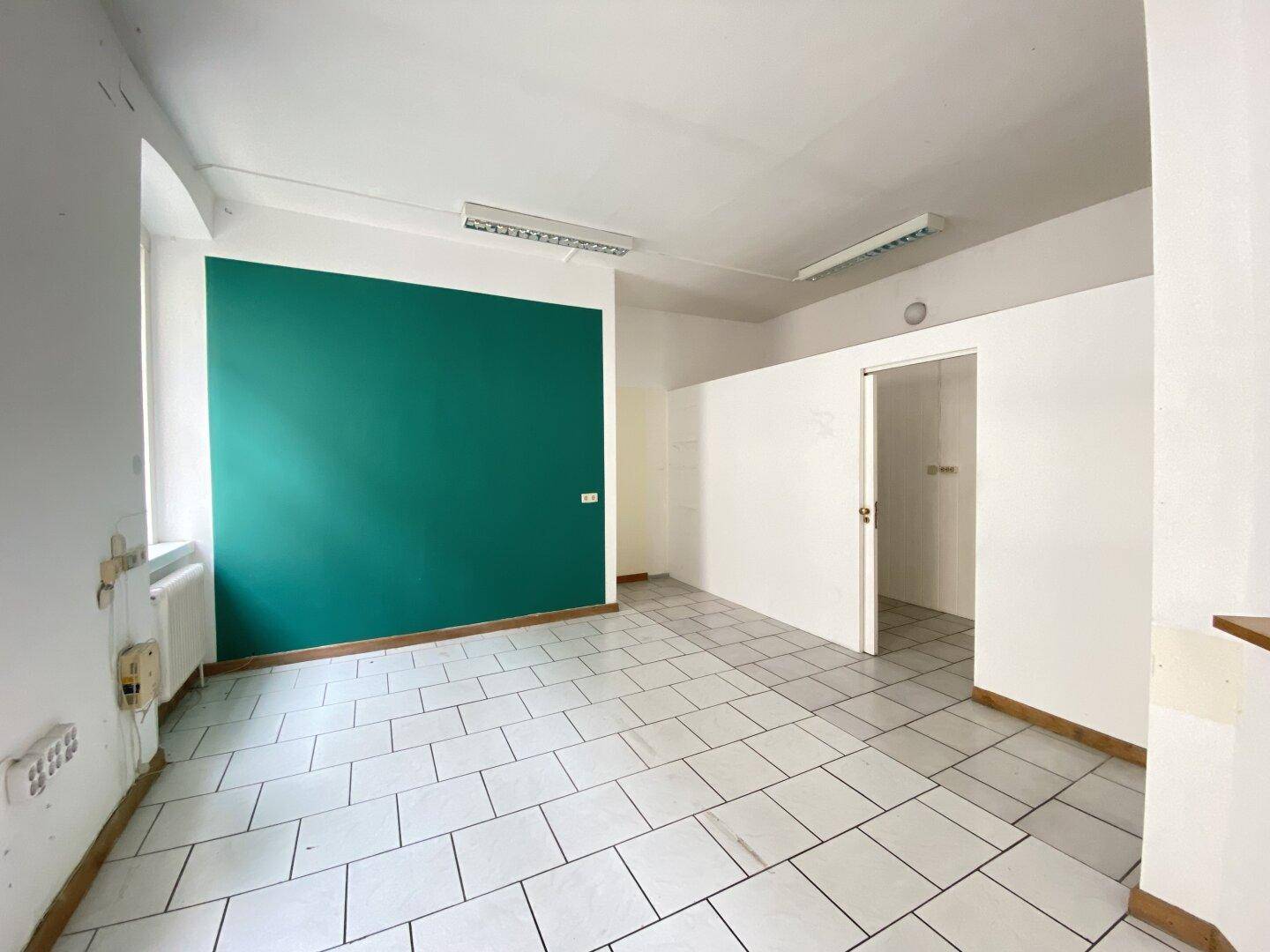 Heller Büroraum mit weißem Fliesenboden, einer tealgrünen Wand und einem offenen Durchgang zu einem Nebenraum.