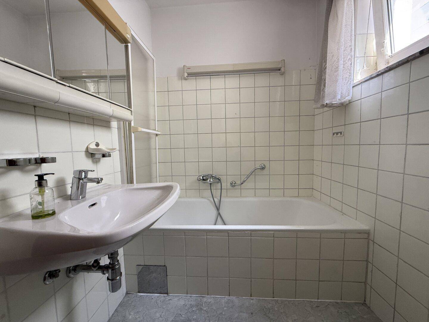 Helles Badezimmer mit Badewanne, Waschbecken, Spiegelschrank und Fenster für Tageslicht.