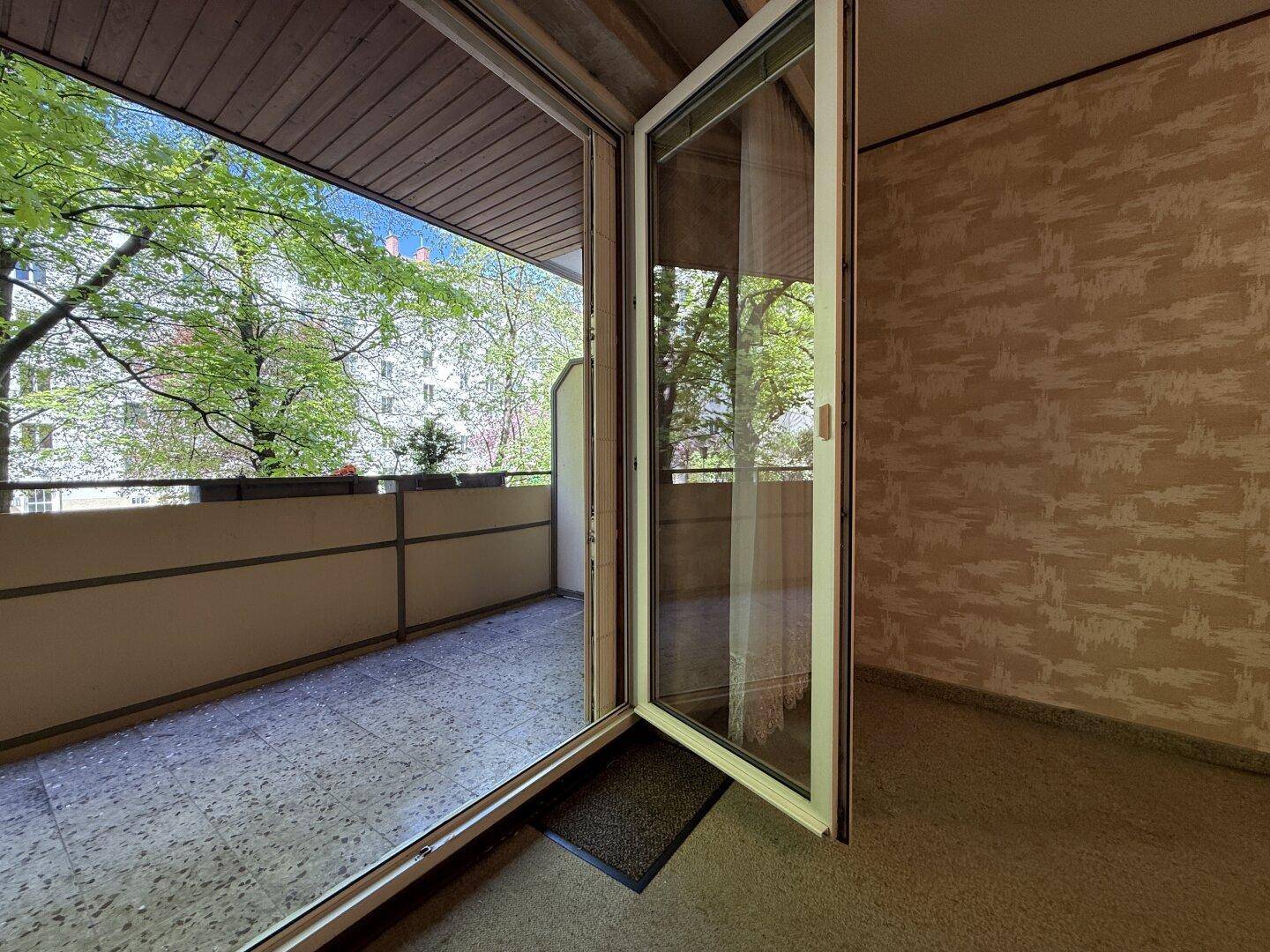 Blick aus einem Innenraum auf einen Balkon mit grünen Bäumen und einfacher Brüstung.