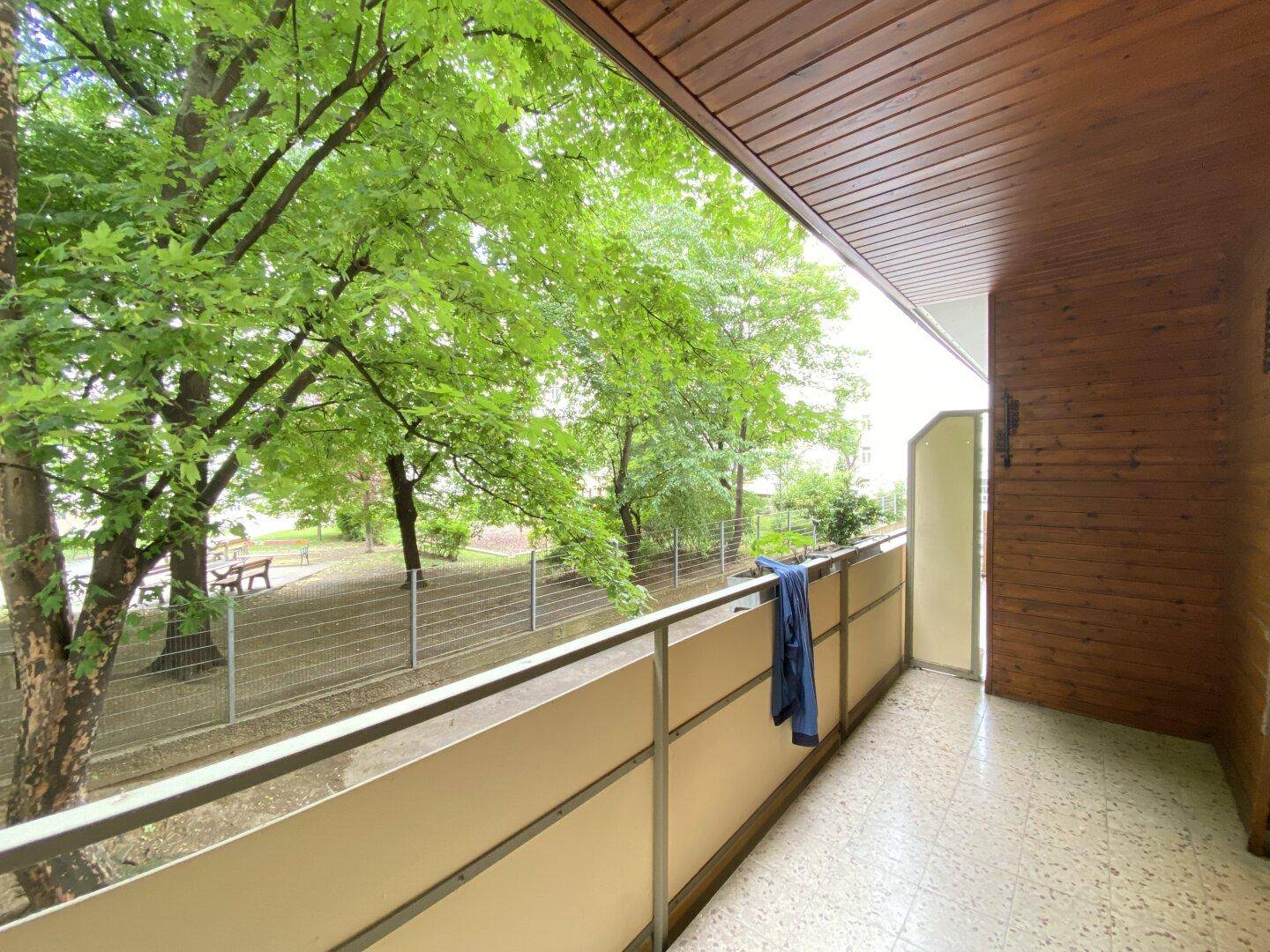 Geräumiger Balkon mit Holzverkleidung und Fliesenboden, bietet einen schönen Blick ins Grüne auf einen Park mit Bänken.