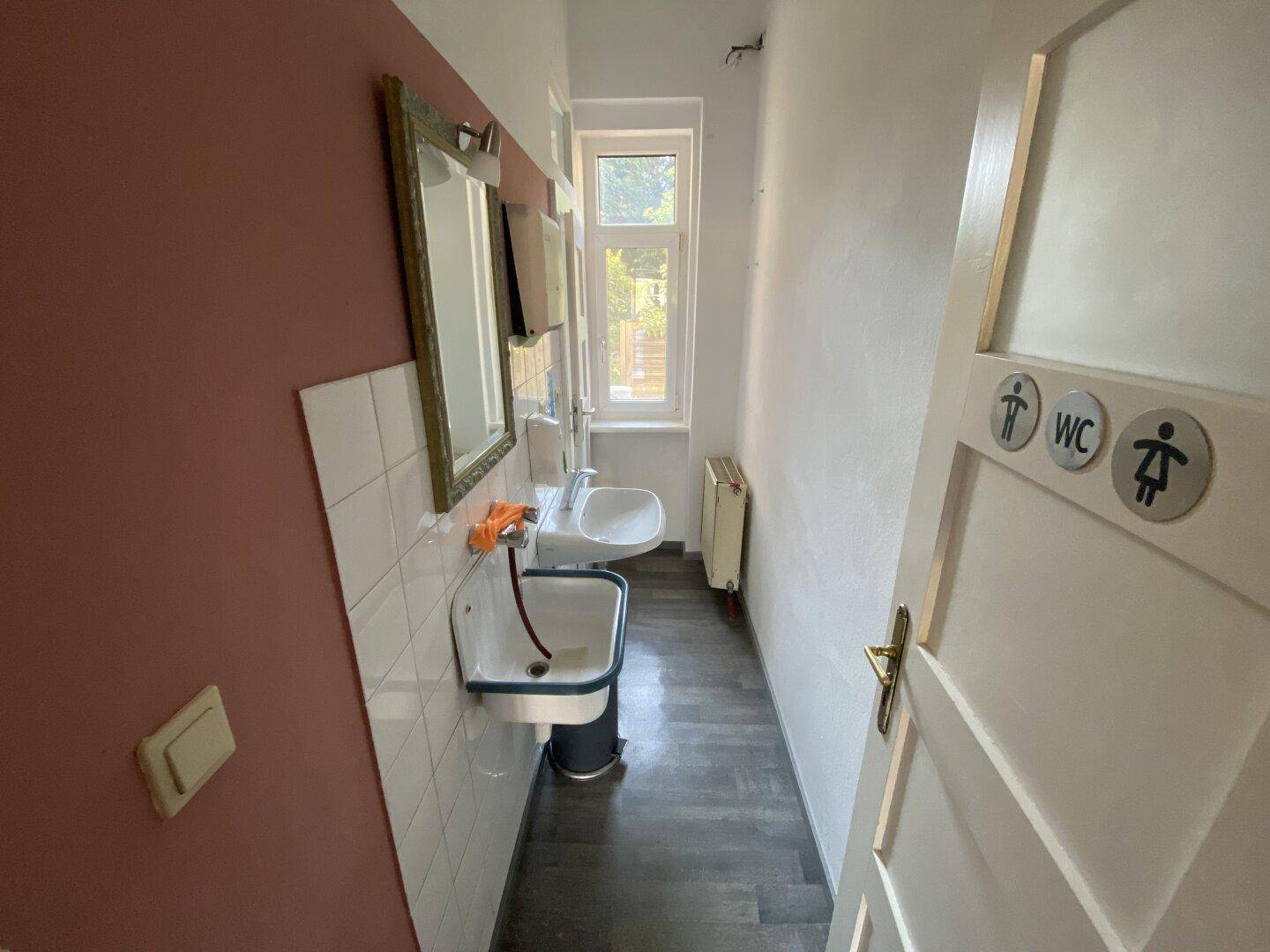 Schmaler Waschraum mit zwei Waschbecken, Fenster und dunklem Boden.