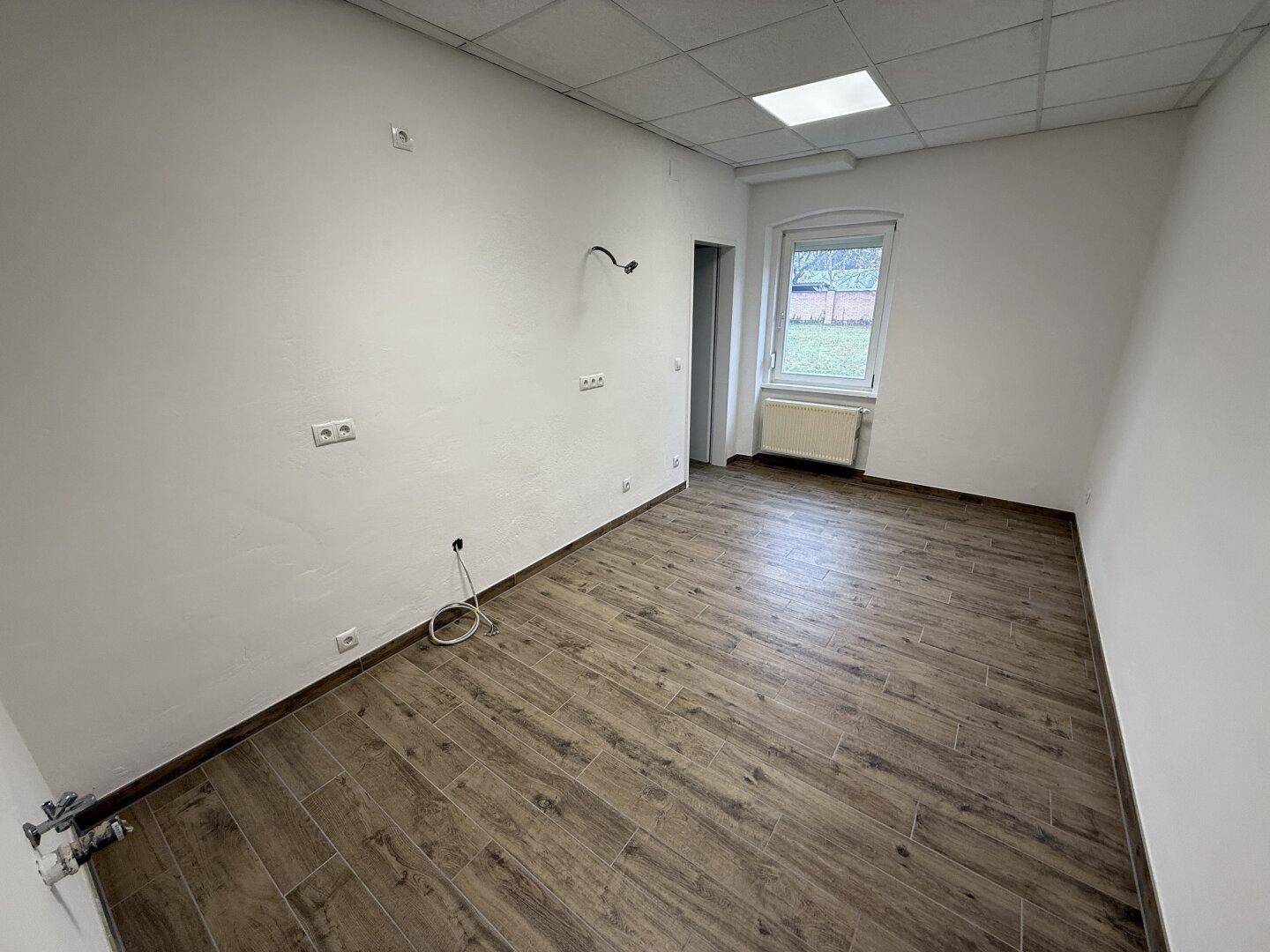 Kleinerer Raum mit Laminatboden, einem Fenster und Anschlüssen an der Wand, möglicherweise für eine Teeküche.