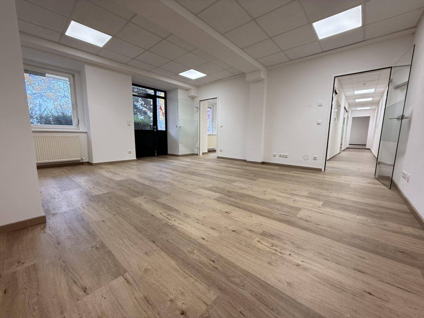Geräumiger, leerer Büroraum mit modernem Laminatboden und großen Fenstern, die viel Tageslicht hereinlassen.