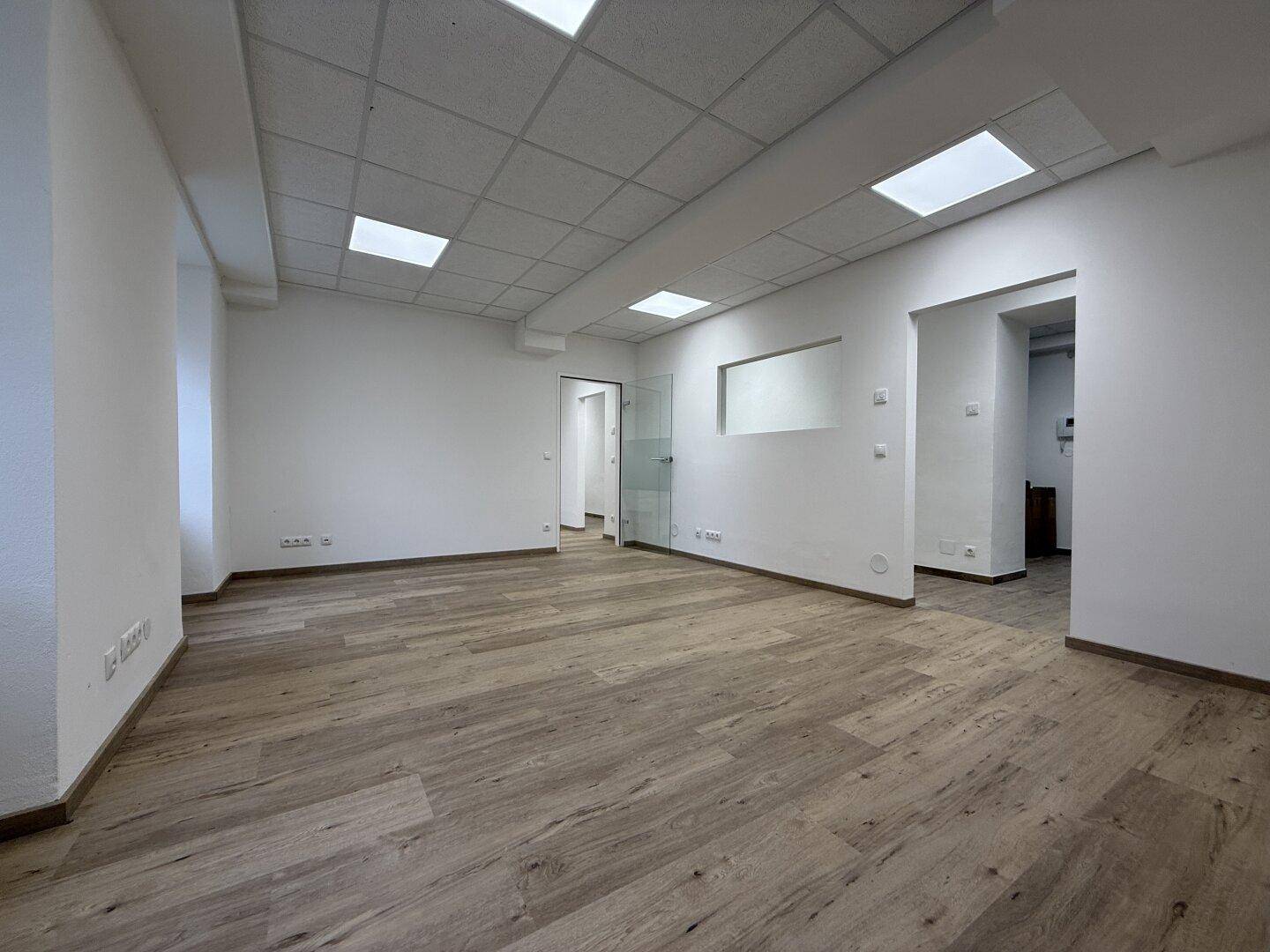 Heller Büroraum mit Laminatboden, abgehängter Decke und Blick auf einen Flur durch eine Glastür.