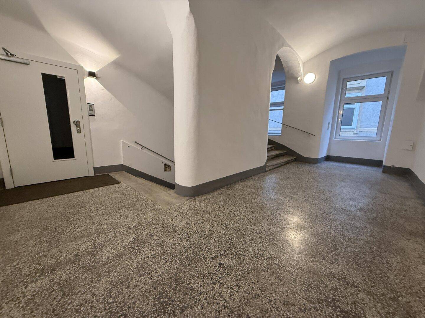 Heller Eingangsbereich mit Terrazzoboden, Treppe und modernem Aufzug, der zu den oberen Etagen führt.