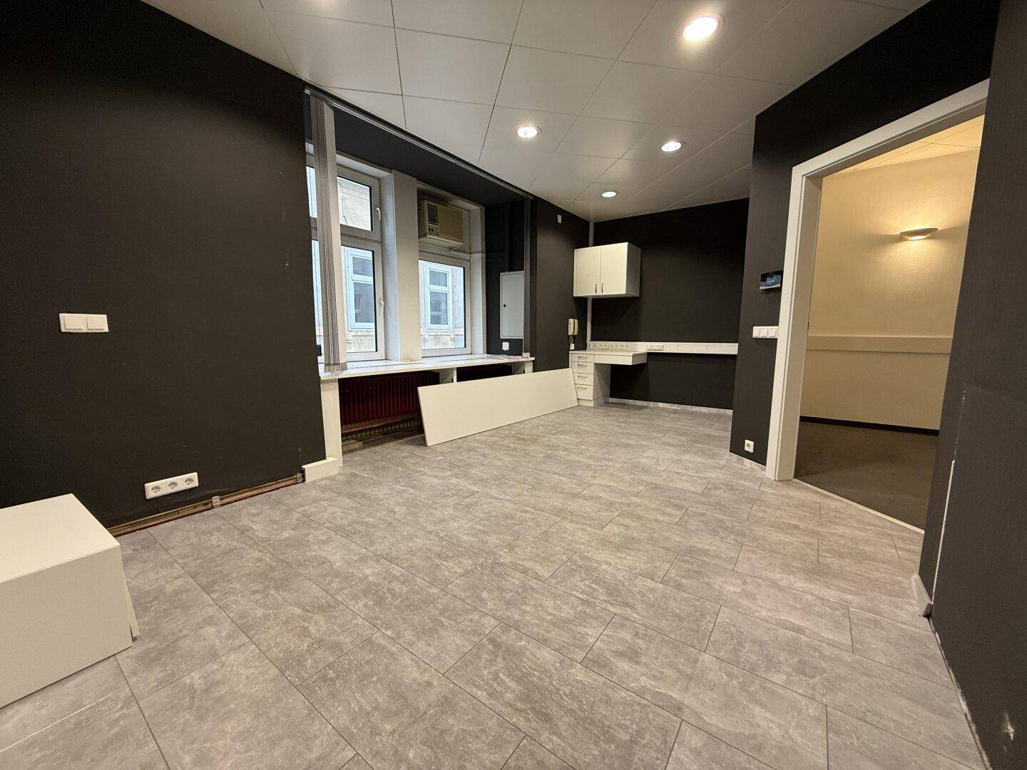 Büroraum mit dunklen Wänden, Fliesenboden und großen Fenstern, teilweise mit Einbauten und Deckenbeleuchtung.