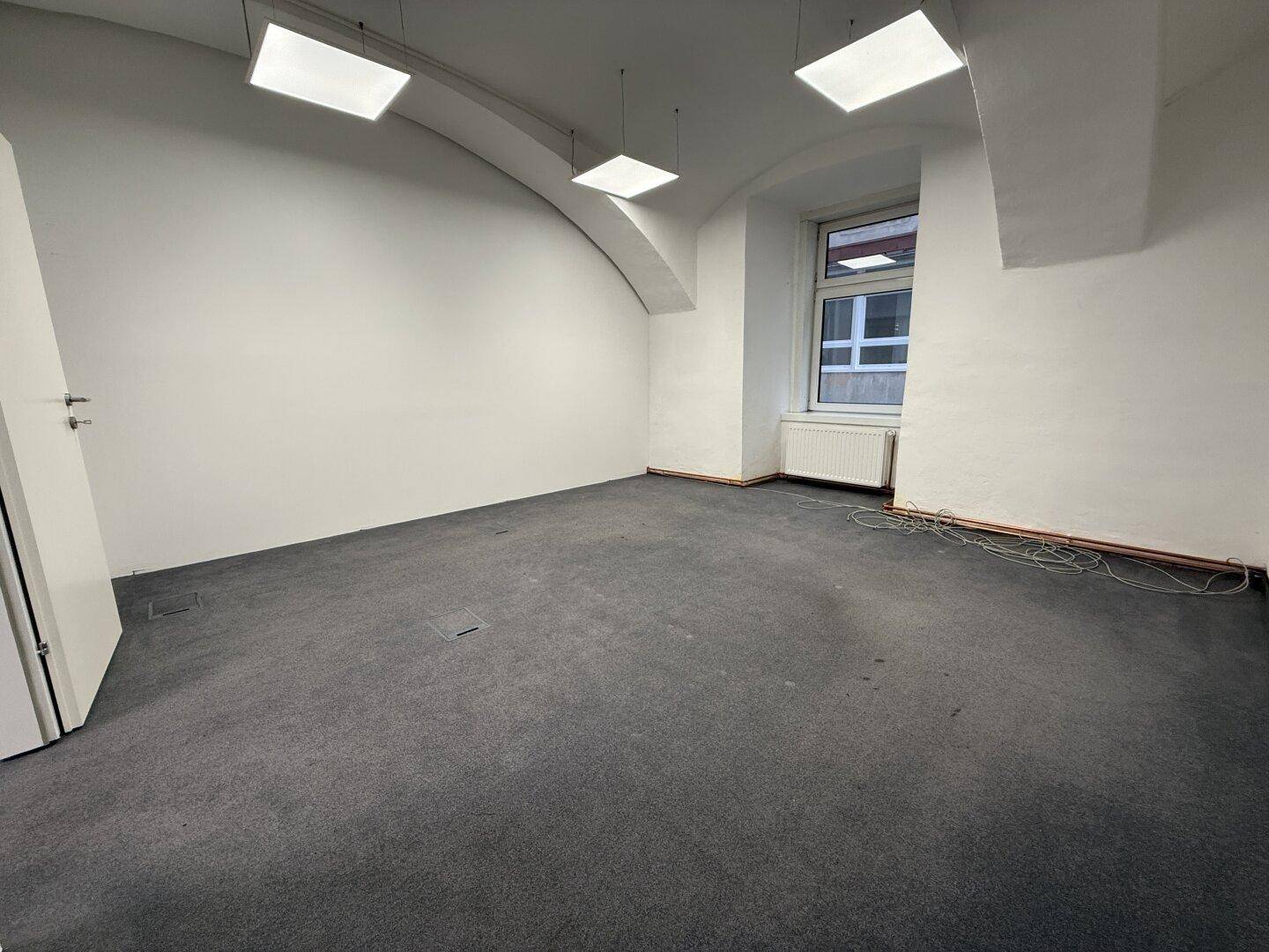 Heller Büroraum mit grauem Teppichboden, einem Fenster und modernen Deckenleuchten, sauber und leer.