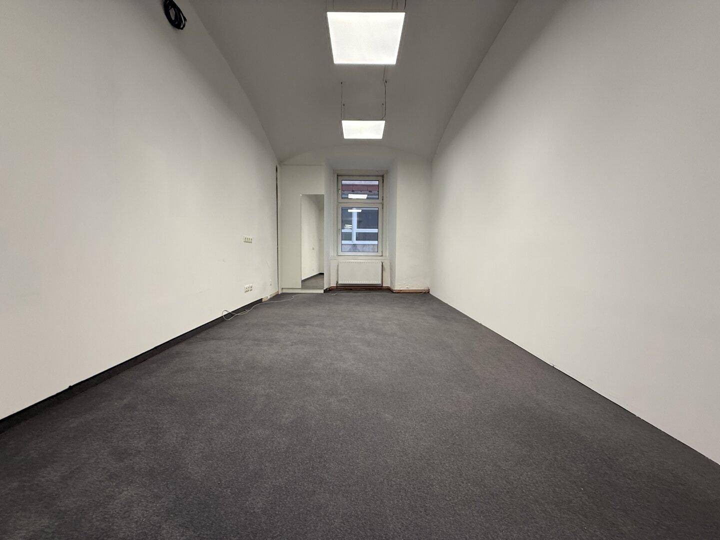 Leerer Büroraum mit grauem Teppichboden und zwei modernen Deckenleuchten, bereit zur Einrichtung und Nutzung.