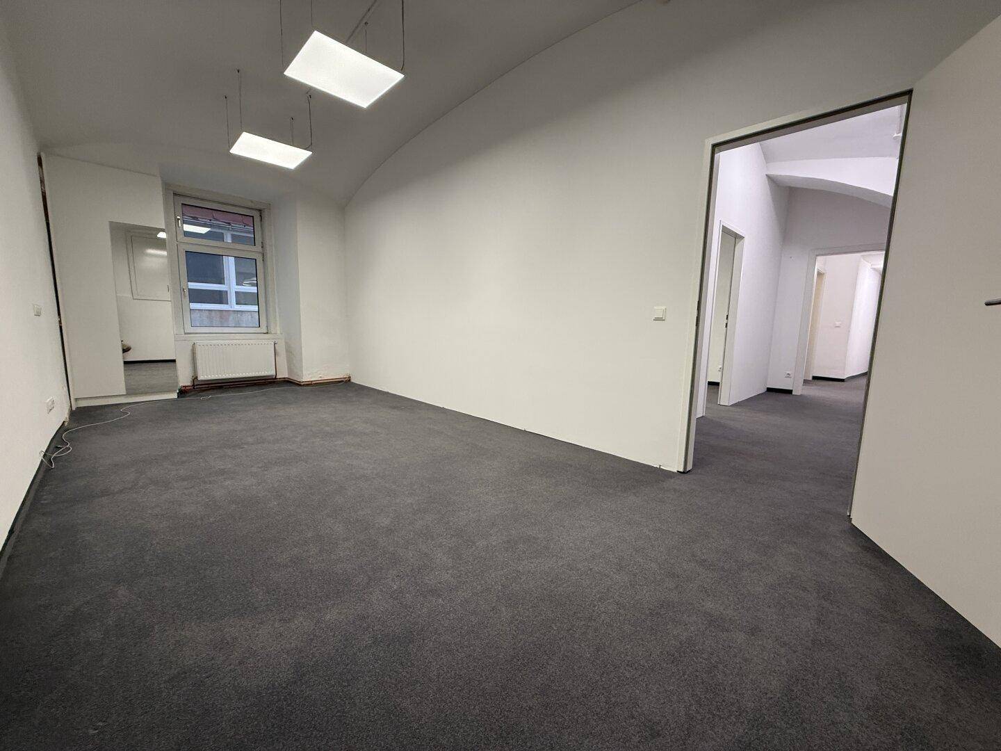 Geräumiger Büroraum mit Teppichboden und Zugang zu weiteren Räumen, hell durch Deckenleuchten und Fenster.