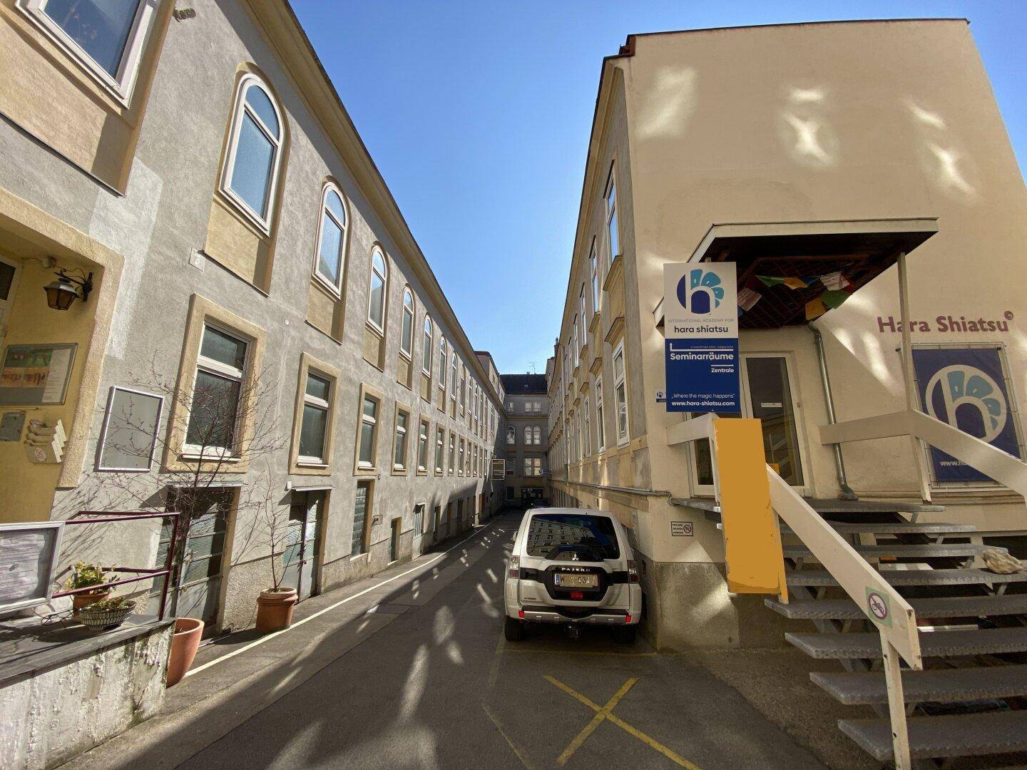 Schmaler Innenhof zwischen zwei Gebäuden mit geparktem Auto und Zugang zu Seminarräumen unter blauem Himmel.