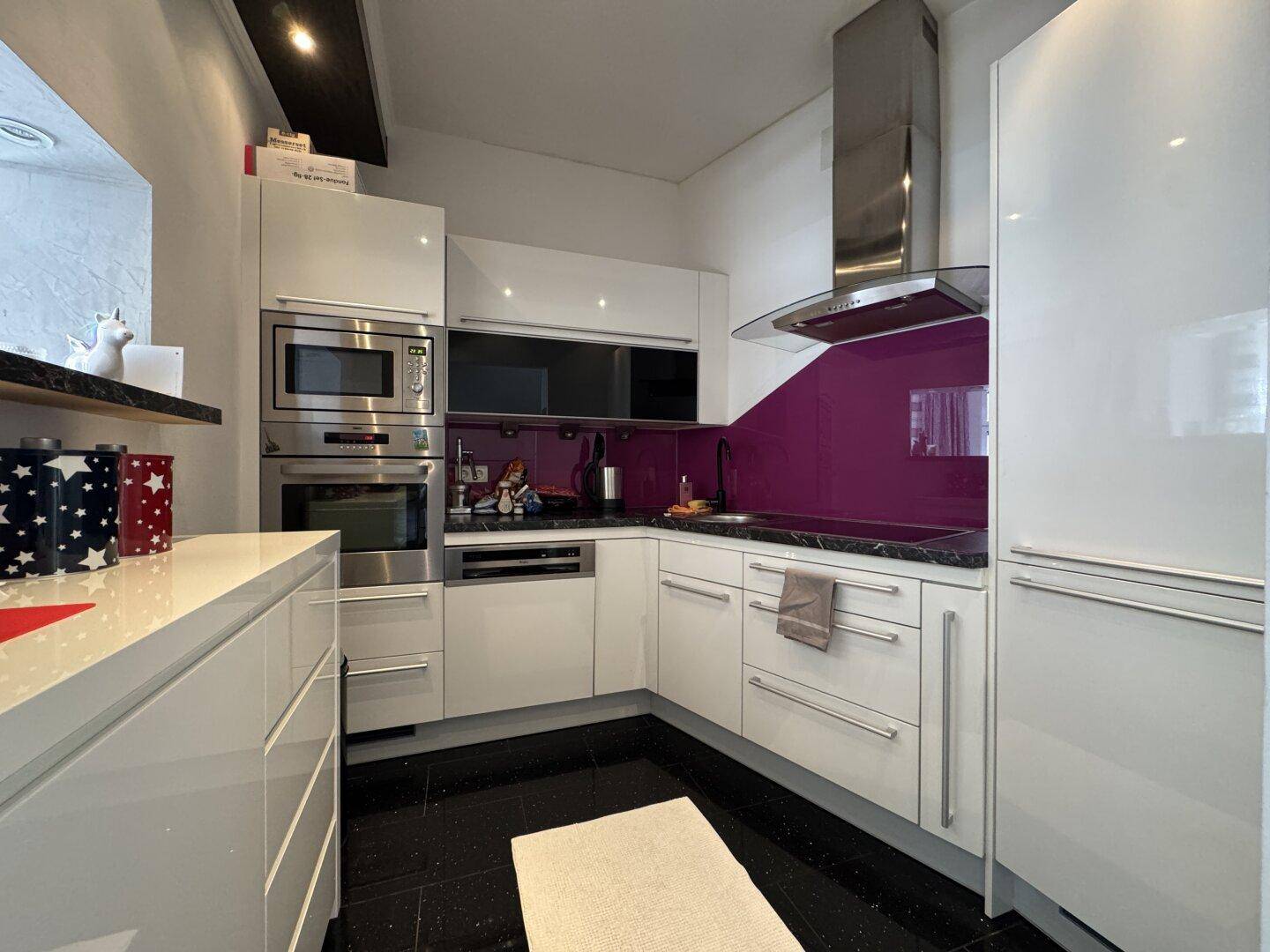 Moderne Einbauküche mit weißen Hochglanzfronten, lila Rückwand und Edelstahlgeräten.