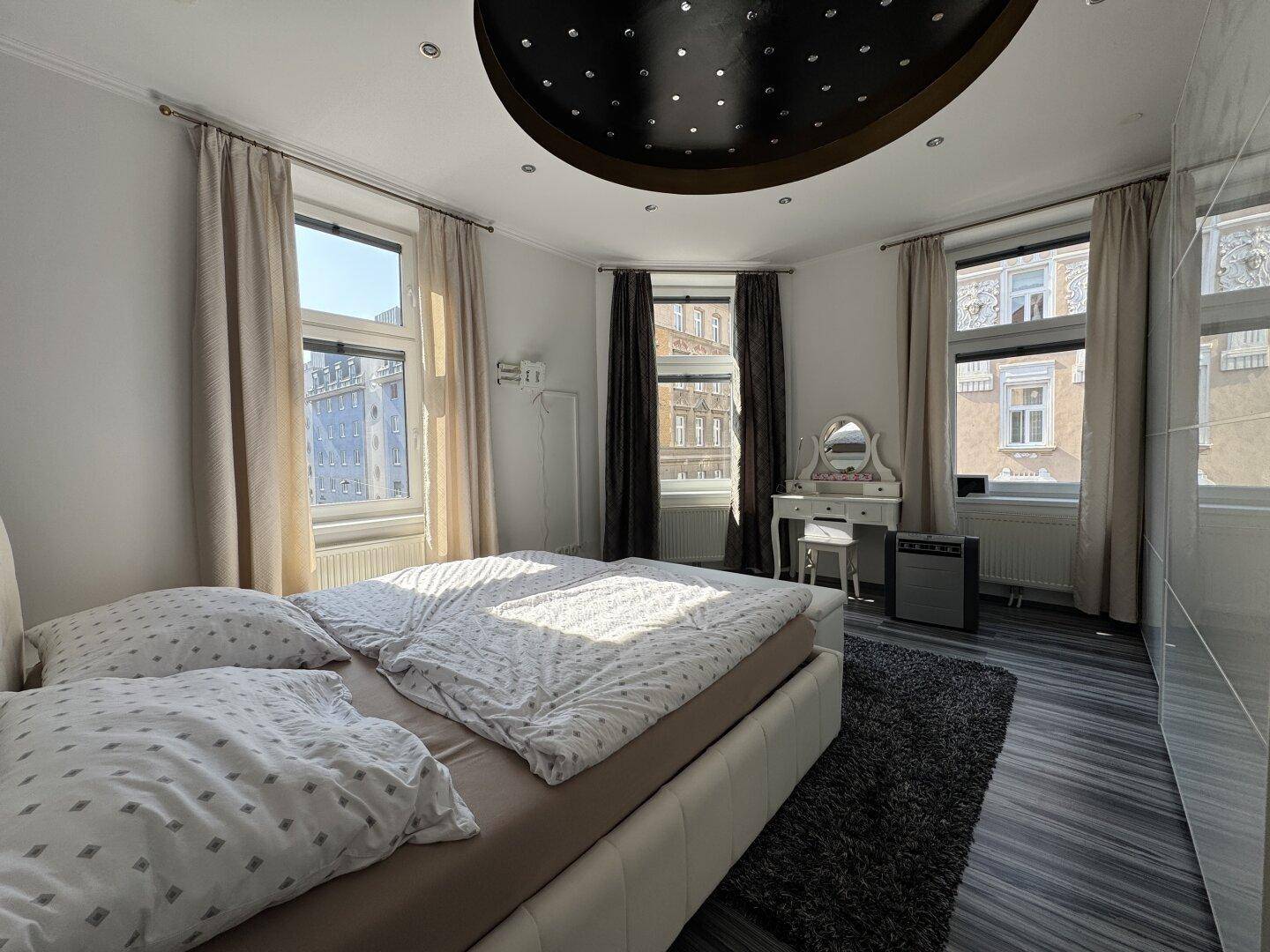 Helles Schlafzimmer mit Doppelbett, dunklem Deckenakzent und Blick auf die umliegenden Gebäude.