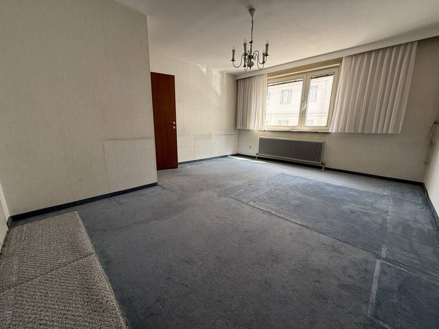 Leerer Raum mit altem blauem Teppichboden und Fenster, bereit für eine Renovierung.