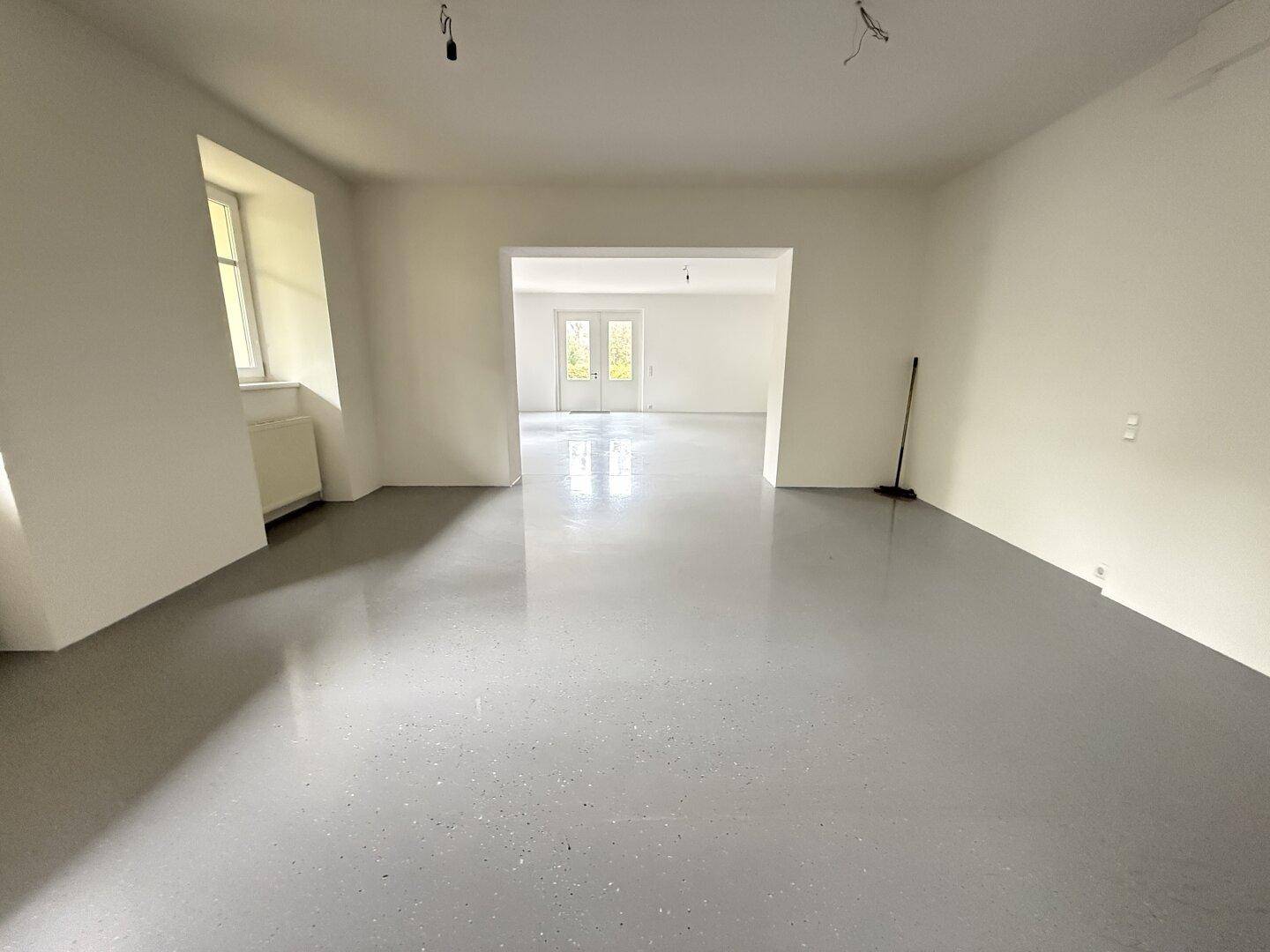 Weitläufiger, heller Raum mit glänzendem Boden und einem Durchgang zu einem weiteren Zimmer mit Fenstertüren.