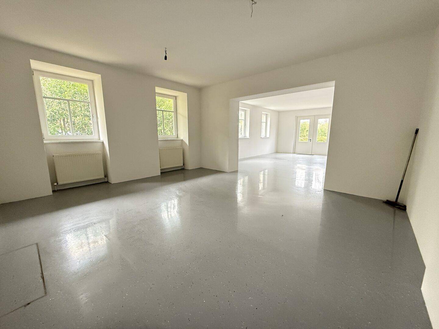 Großer, heller Raum mit mehreren Fenstern und einem Durchgang zu einem weiteren Bereich mit Fenstertüren.