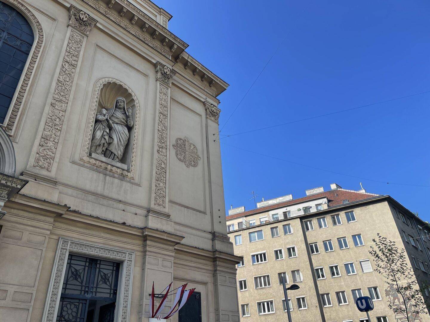 Fassade eines historischen Gebäudes mit Skulptur und angrenzendem modernen Wohnhaus unter blauem Himmel.
