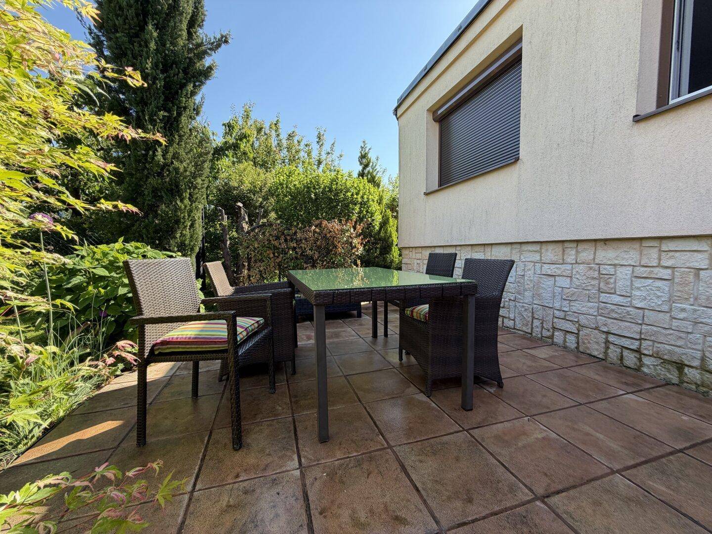 Geflieste Terrasse mit Esstisch und Stühlen, umgeben von üppiger Bepflanzung und einer Steinmauer.