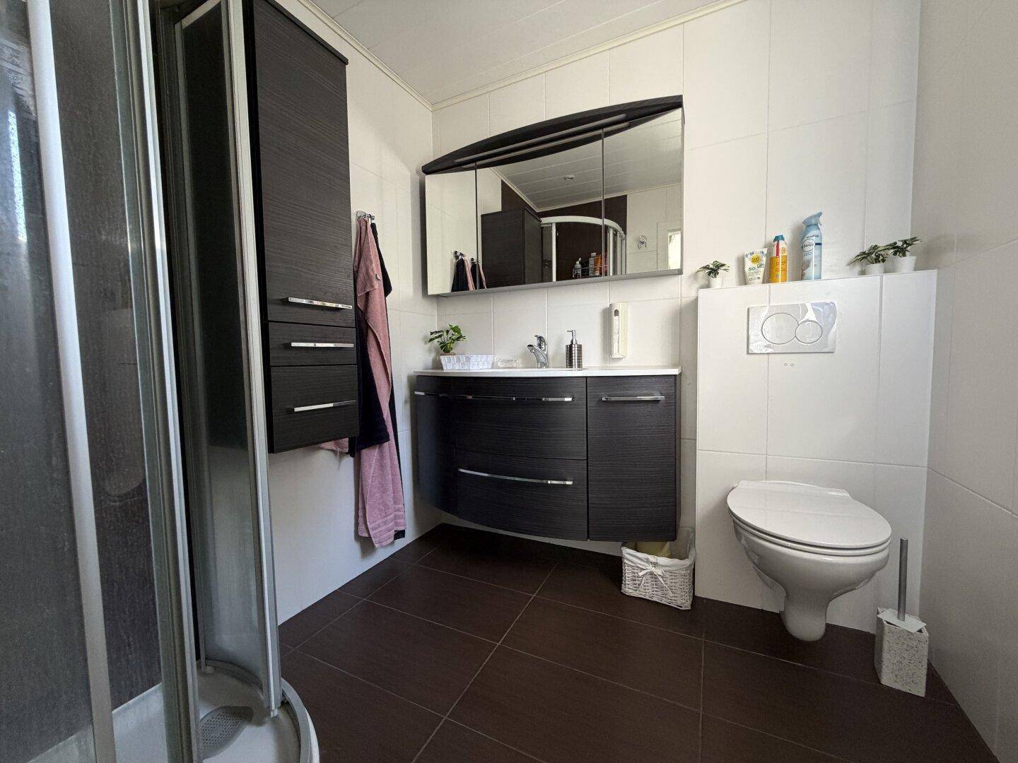 Modernes Badezimmer mit Duschkabine, Hänge-WC, Waschtisch und dunklen Fliesen.