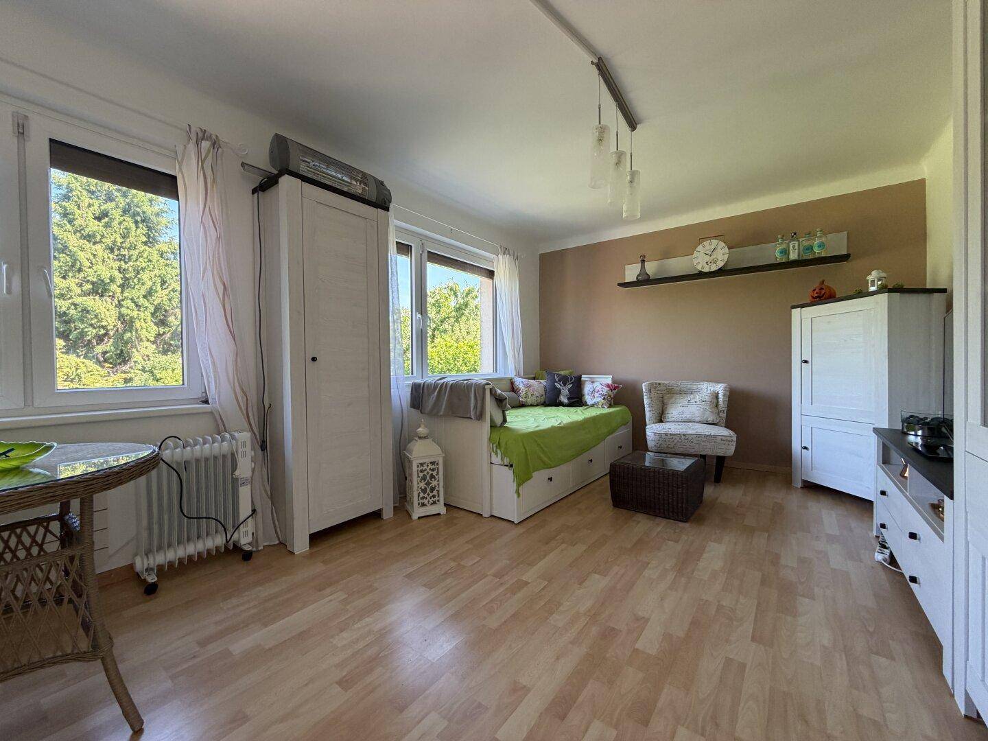 Helles Wohnzimmer mit Laminatboden, großen Fenstern und einfacher Einrichtung.