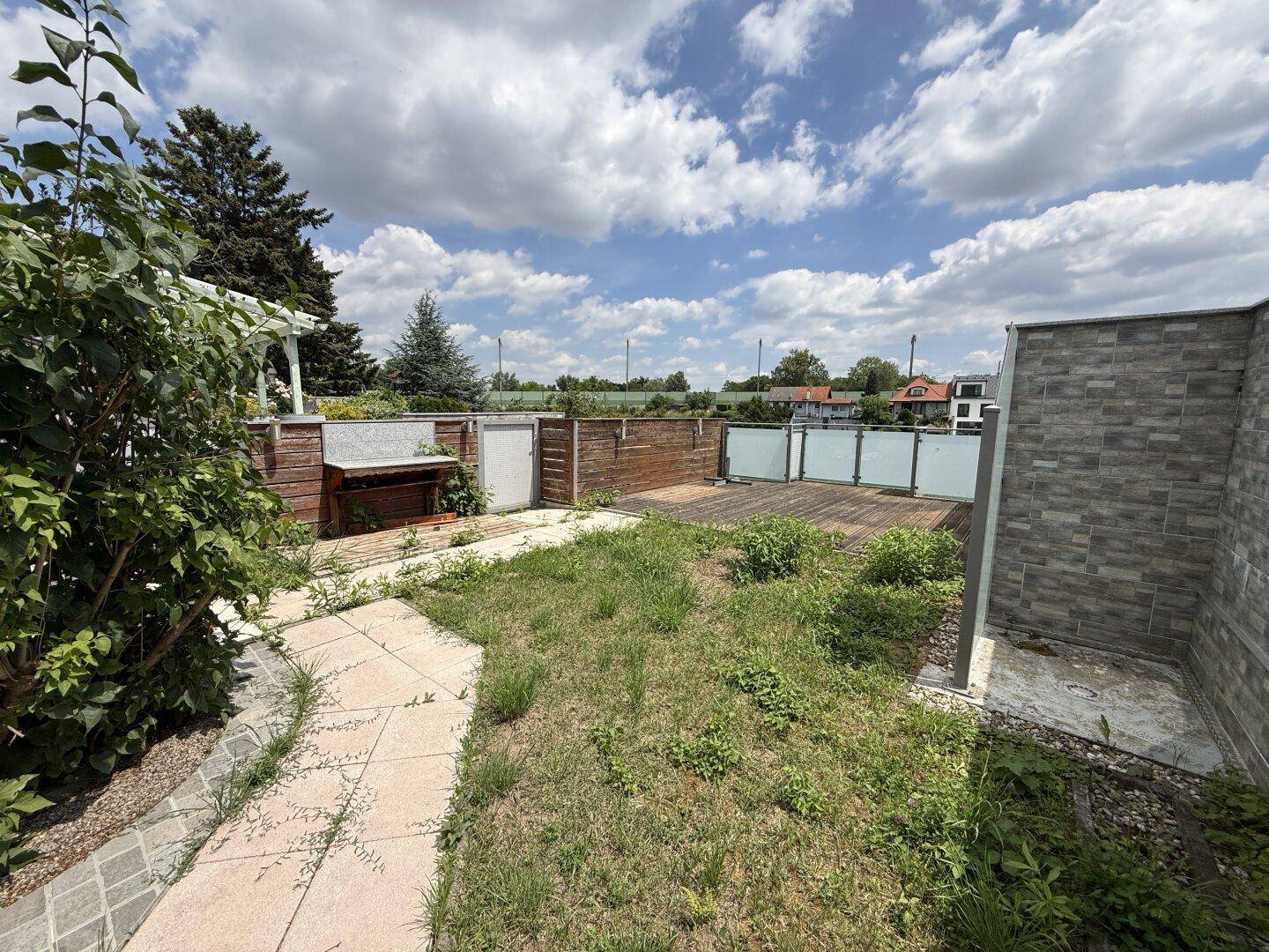 Gartenbereich mit Rasenfläche, gepflastertem Weg und einer Holzterrasse im Hintergrund.