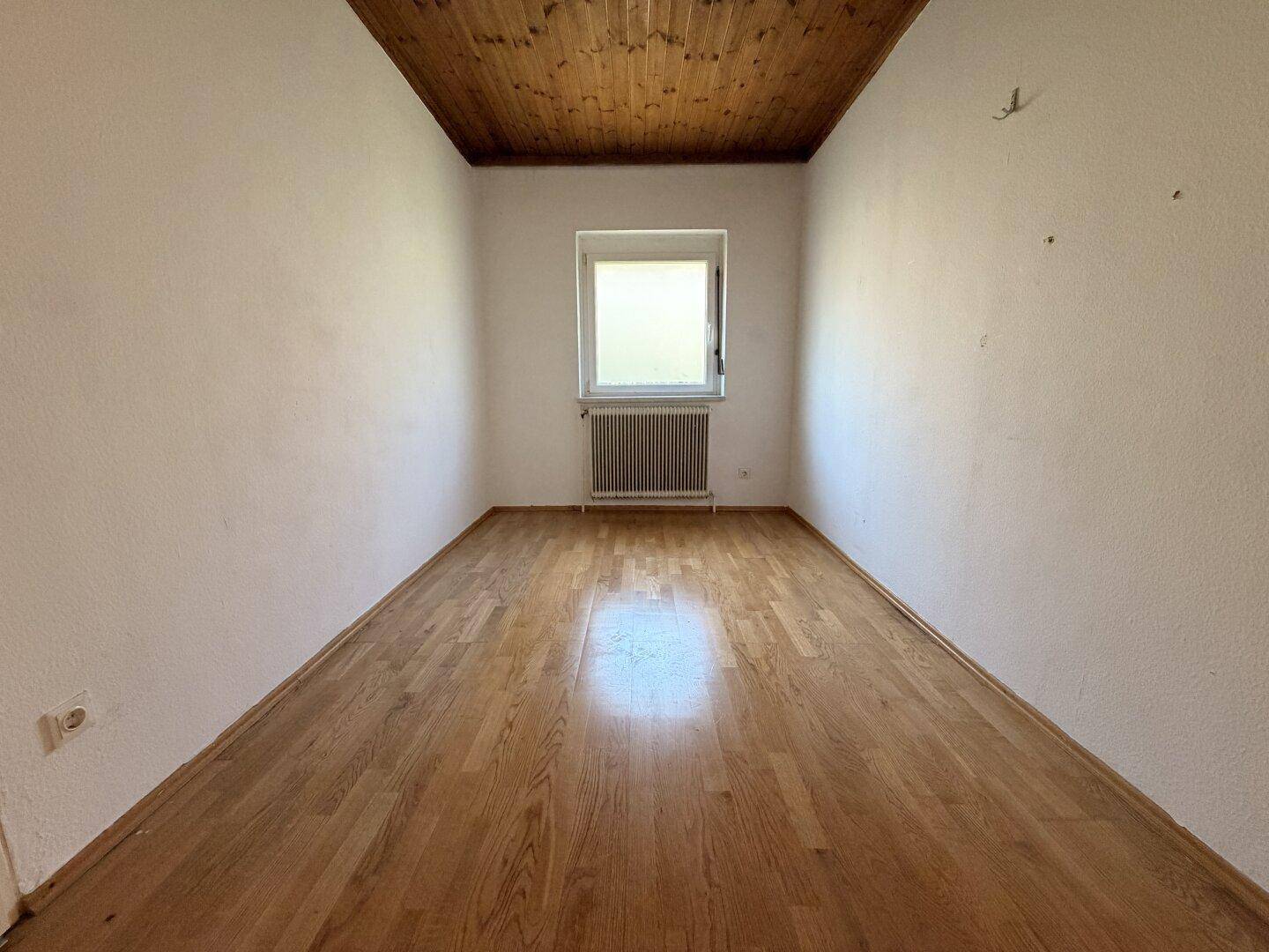 Helles Zimmer mit Holzbalkendecke, Parkettboden und einem Fenster mit Blick ins Grüne.