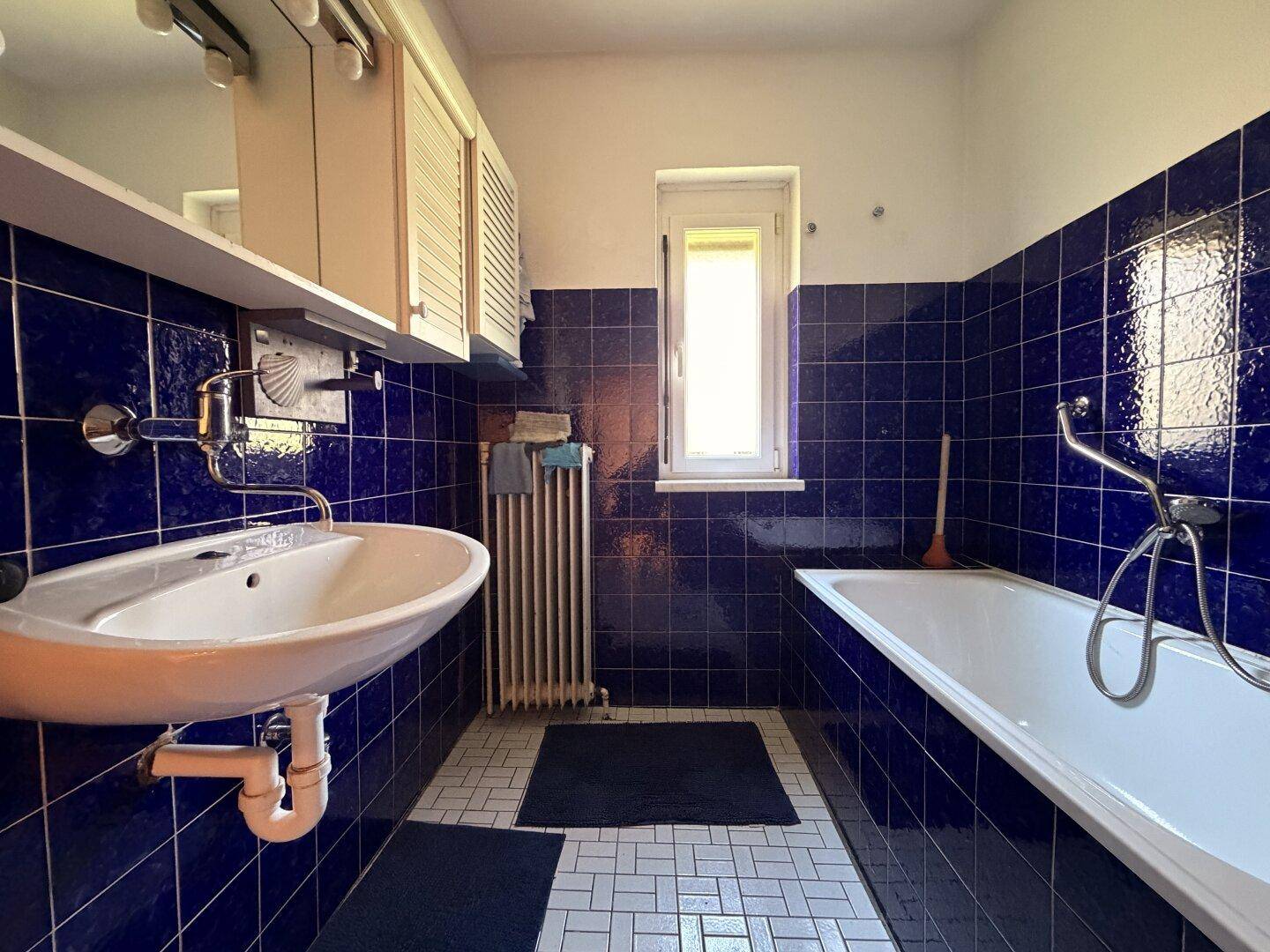 Badezimmer mit blauen Fliesen, Badewanne, Waschbecken und einem Fenster für Tageslicht.