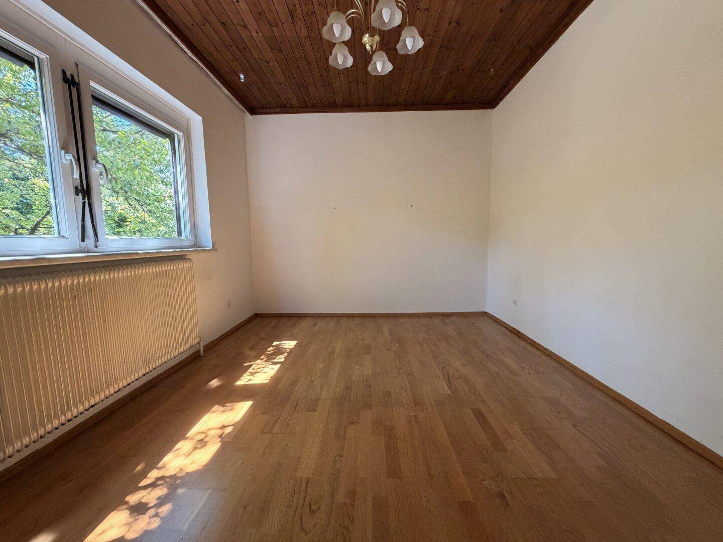Sonniges Zimmer mit Holzbalkendecke, Parkettboden und großem Fenster mit Blick nach draußen.