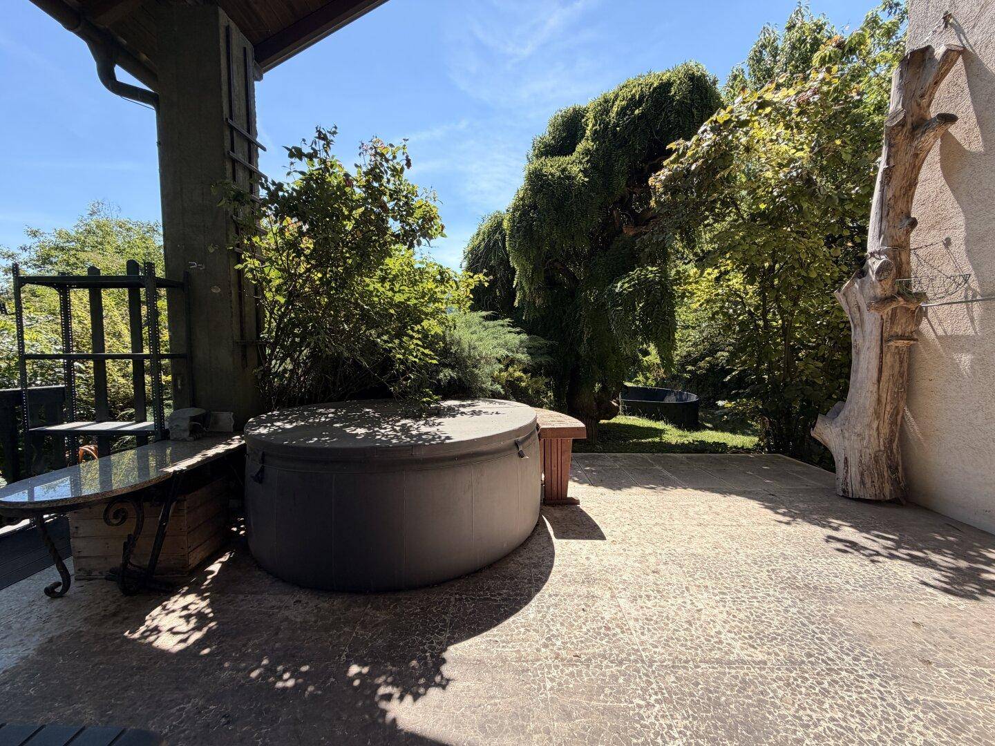 Geräumige Terrasse mit Whirlpool und Holztisch, umgeben von üppiger Vegetation und Bäumen.
