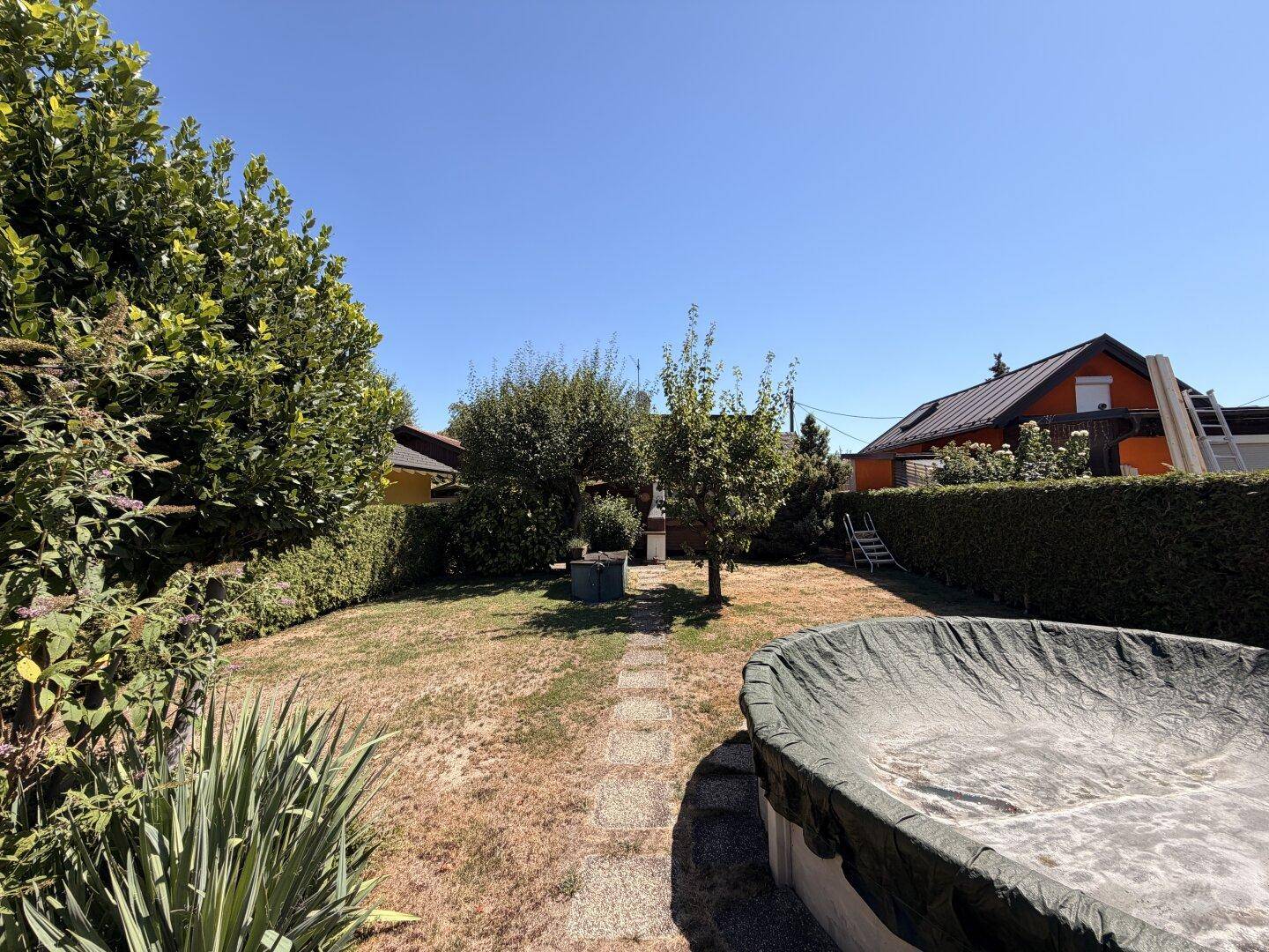 Weitläufiger Garten mit trockenem Rasen, üppigen Hecken, Bäumen und einem abgedeckten Aufstellpool im Sonnenlicht.