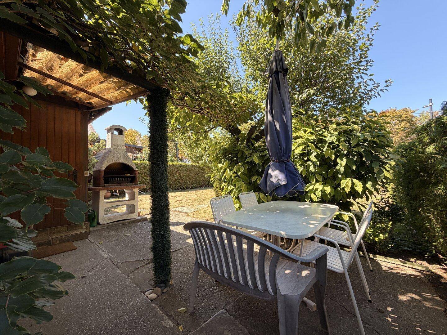 Gemütlicher Sitzbereich auf der Terrasse unter einer Pergola mit Gartenmöbeln und Blick auf den Grill.