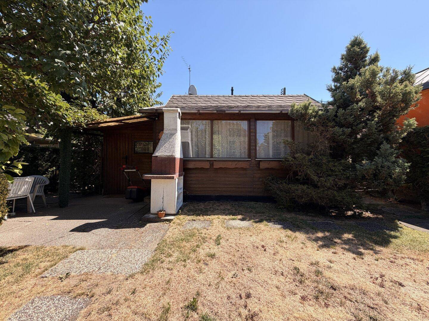 Kleines Holzhaus mit überdachter Terrasse und gemauertem Grill im Gartenbereich.