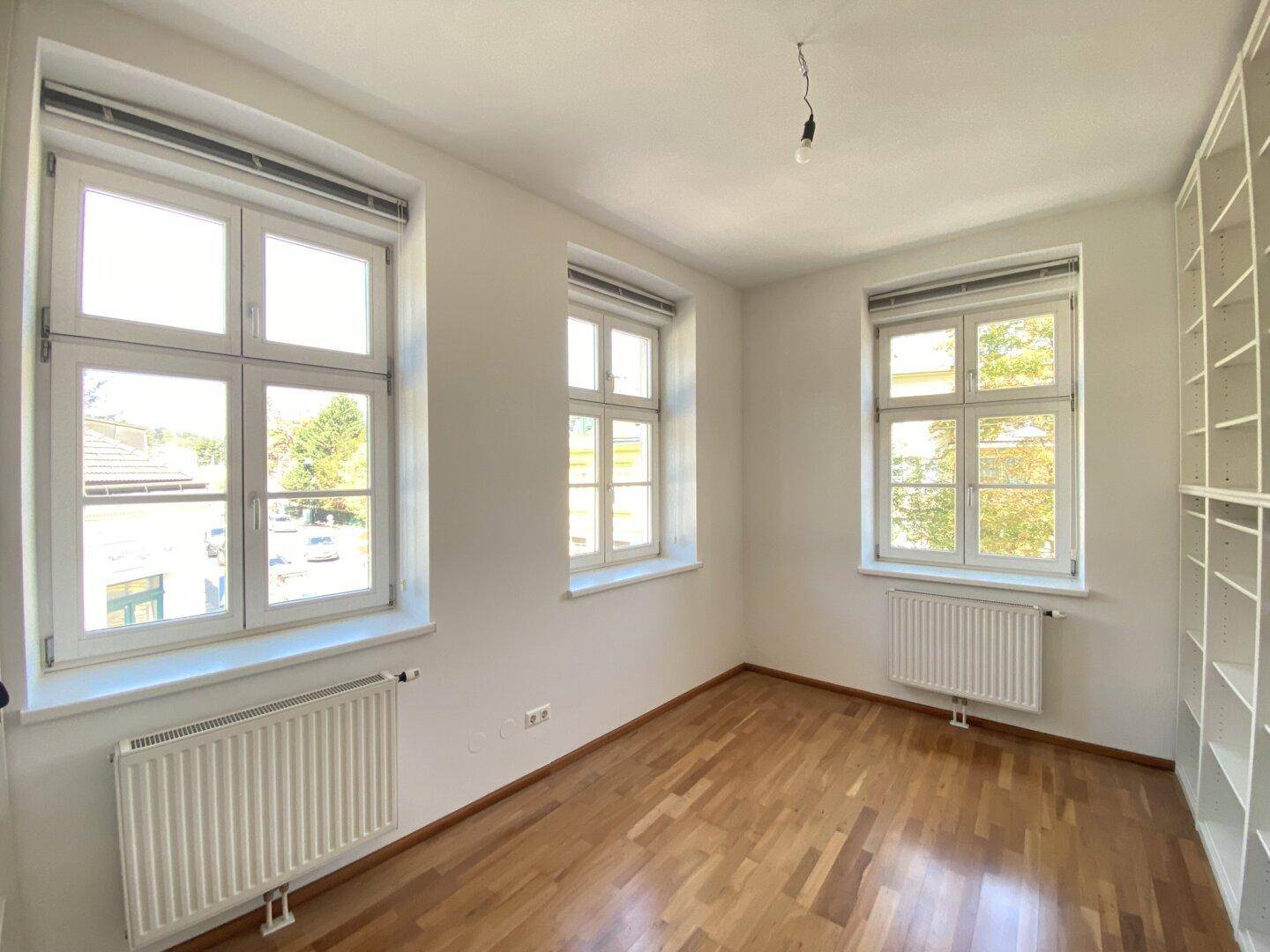Helles Schlafzimmer mit Holzparkettboden, drei Fenstern und eingebauten Regalen an der Wand.