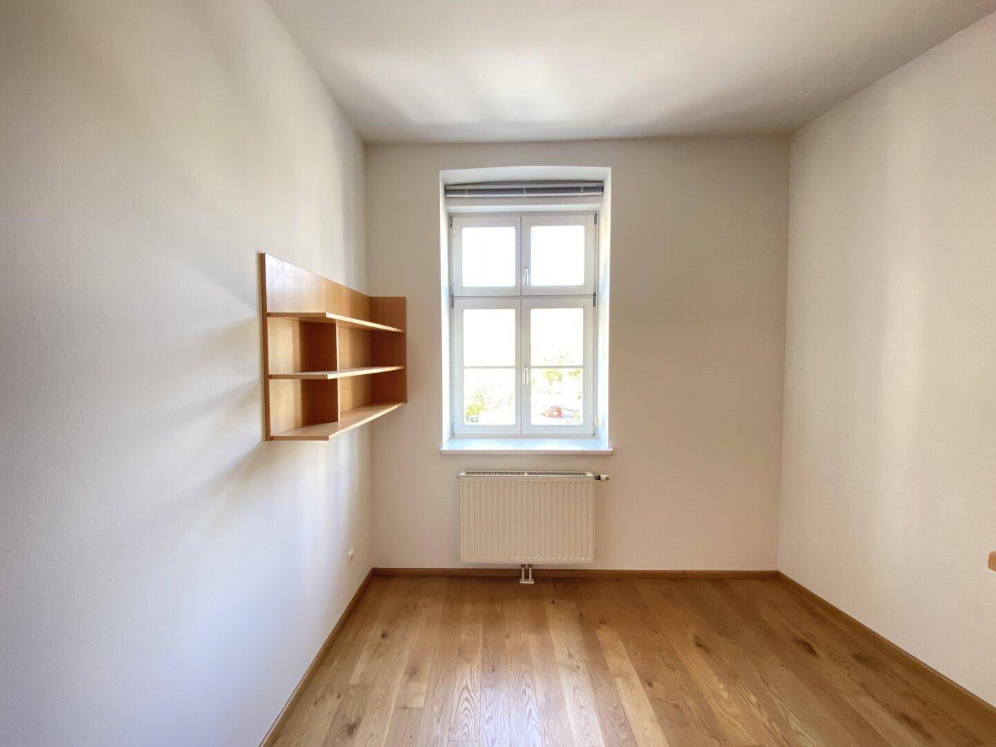 Kleineres Schlafzimmer mit Holzparkettboden, einem Fenster und einer schwebenden Holzablage.