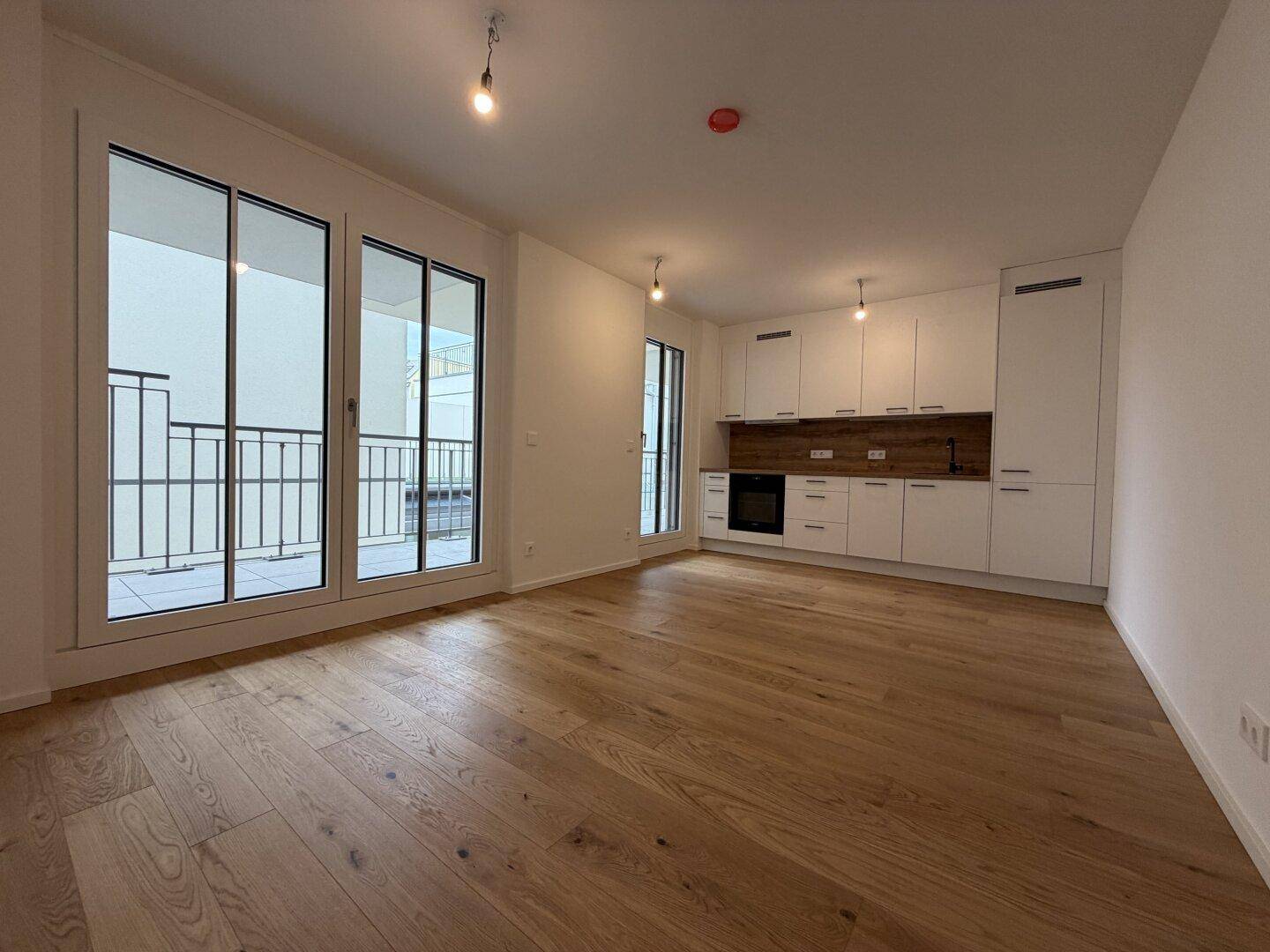 Heller offener Wohnbereich mit moderner Einbauküche, Holzboden und Zugang zum Balkon.