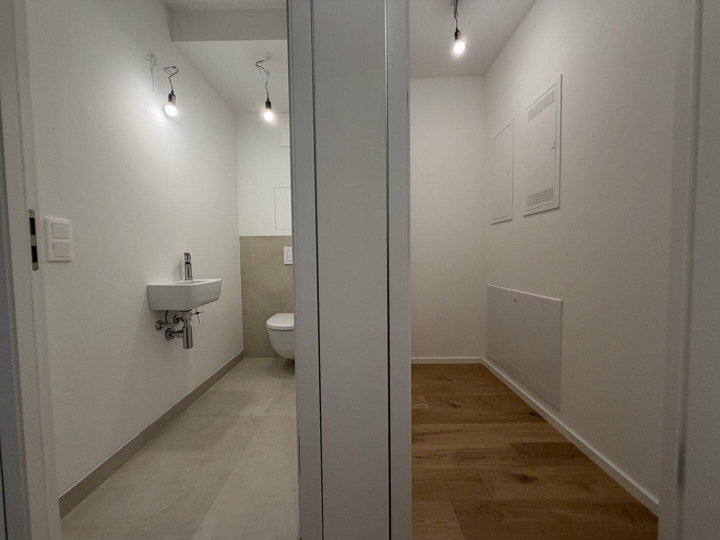 Modernes Gäste-WC mit Wand-WC und kleinem Waschbecken, daneben ein leerer Raum.