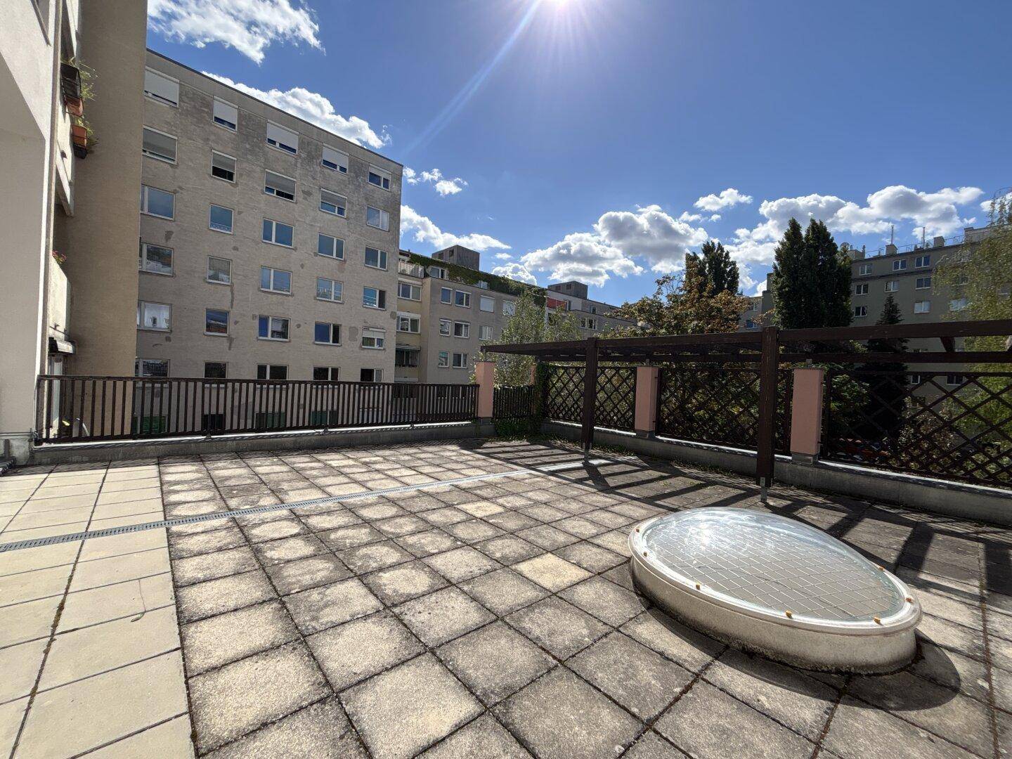 Große, sonnige Dachterrasse mit Steinplatten und Blick auf umliegende Gebäude.