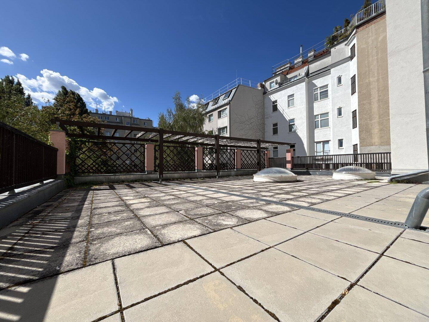 Weitläufige Dachterrasse mit Steinplatten und Pergola, umgeben von anderen Gebäuden.