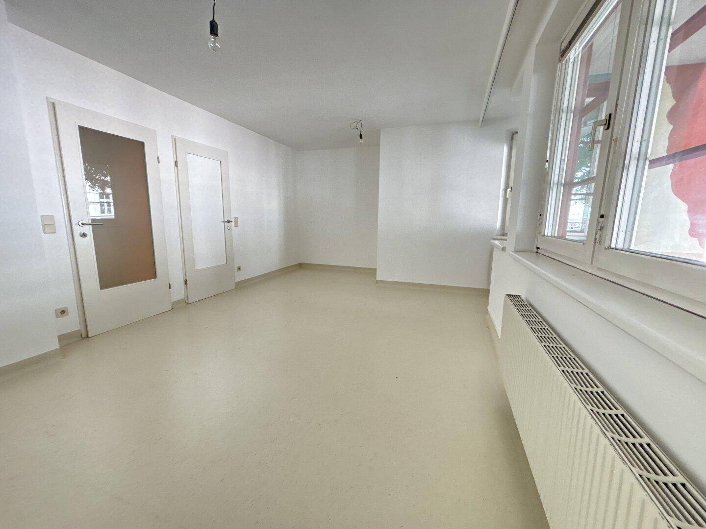 Heller, leerer Raum mit weißen Wänden, großen Fenstern und einem hellen Vinylboden.