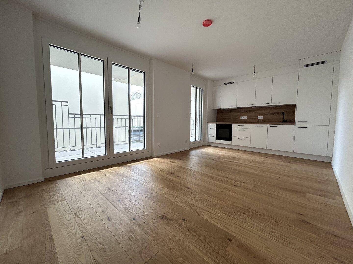 Heller offener Wohnbereich mit integrierter weißer Küche, Holzboden und Zugang zum Balkon.