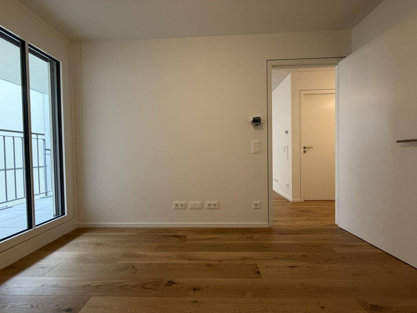 Leeres Zimmer mit Holzboden, weißen Wänden und Zugang zu einem weiteren Raum und Außenbereich.