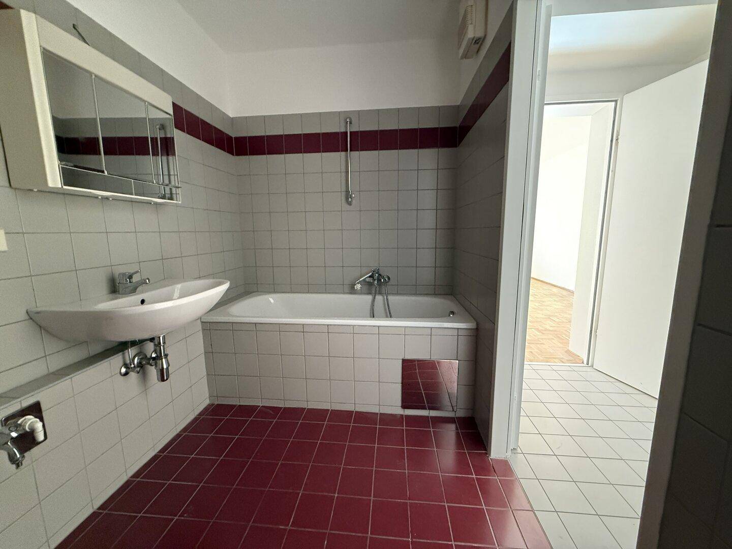 Badezimmer mit Badewanne, Waschbecken und Fliesen in Grau und Rot, mit einem Spiegelschrank.