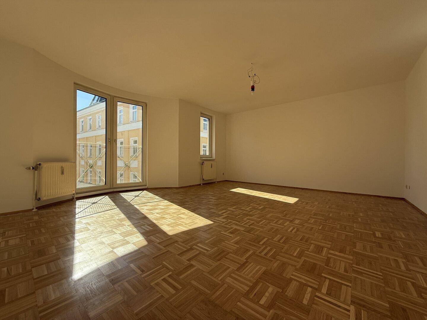 Geräumiger, unmöblierter Wohnbereich mit hellem Parkettboden und Zugang zu einem Balkon, durchflutet von Sonnenlicht.