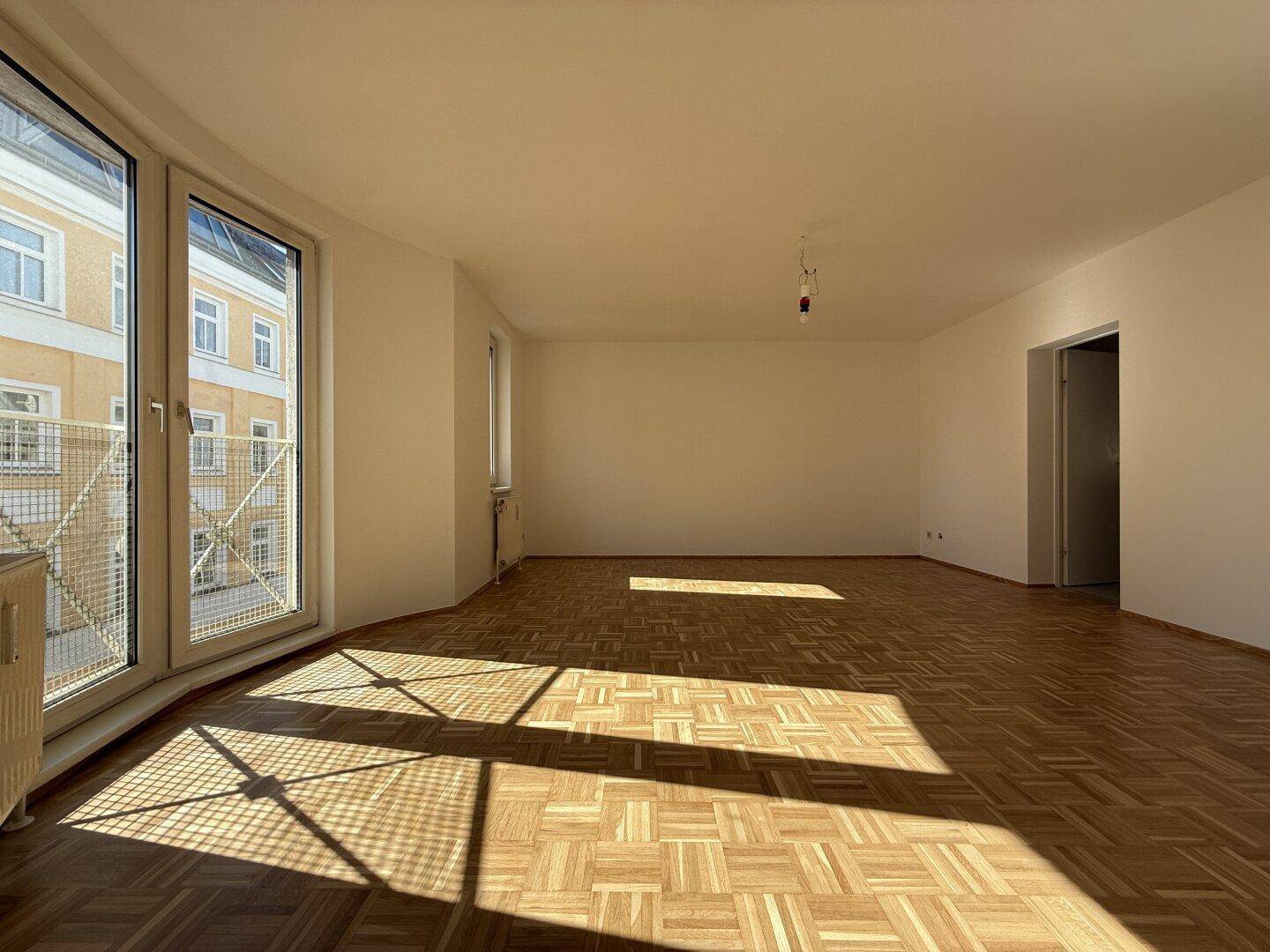 Lichtdurchfluteter Wohnraum mit elegantem Parkettboden und breiten Fenstern, die einen Blick nach draußen bieten.