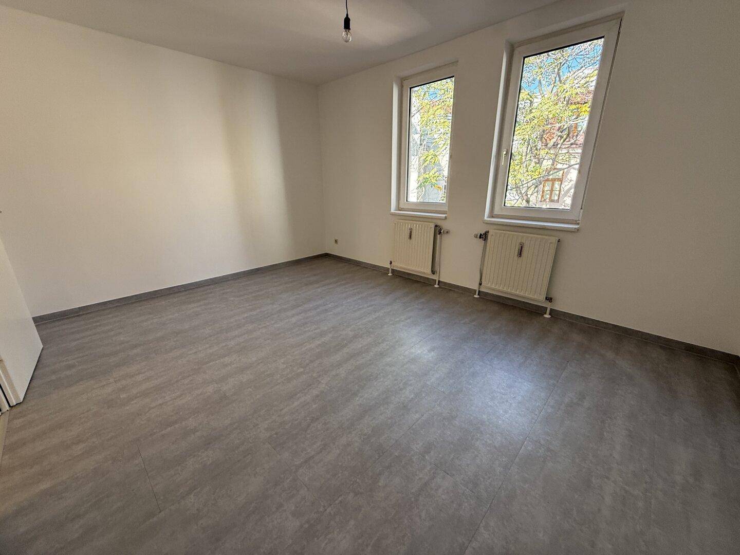 Leeres Zimmer mit modernem grauem Vinylboden und zwei Fenstern, die den Raum mit Tageslicht erhellen.