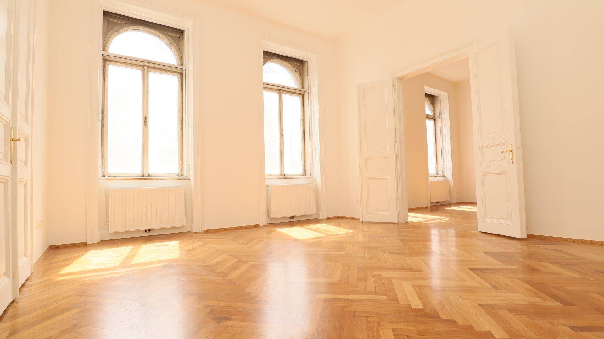 Geräumiges Zimmer mit hellem Fischgrätparkett und hohen Fenstern, das viel Tageslicht hereinlässt.