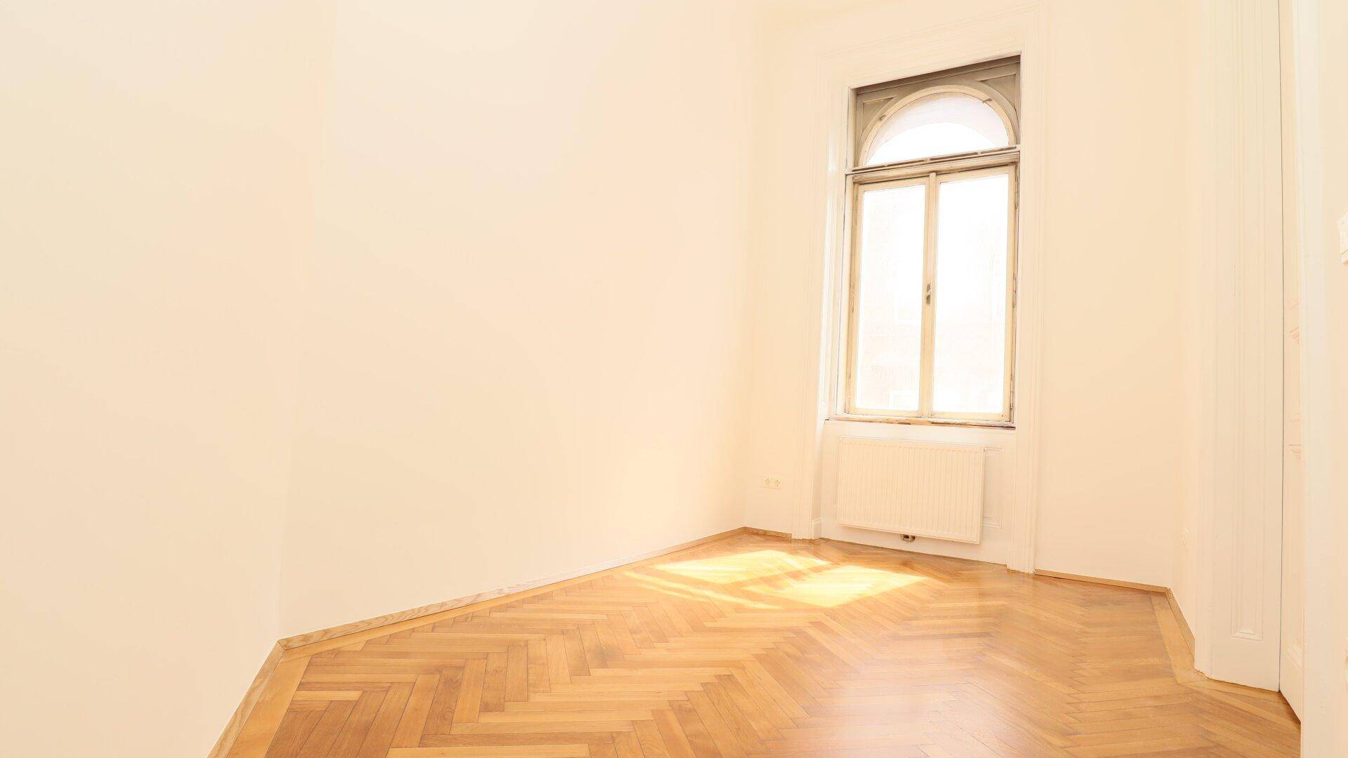 Gemütliches Zimmer mit Fischgrätparkett und einem hohen Bogenfenster, das viel Sonnenlicht empfängt.