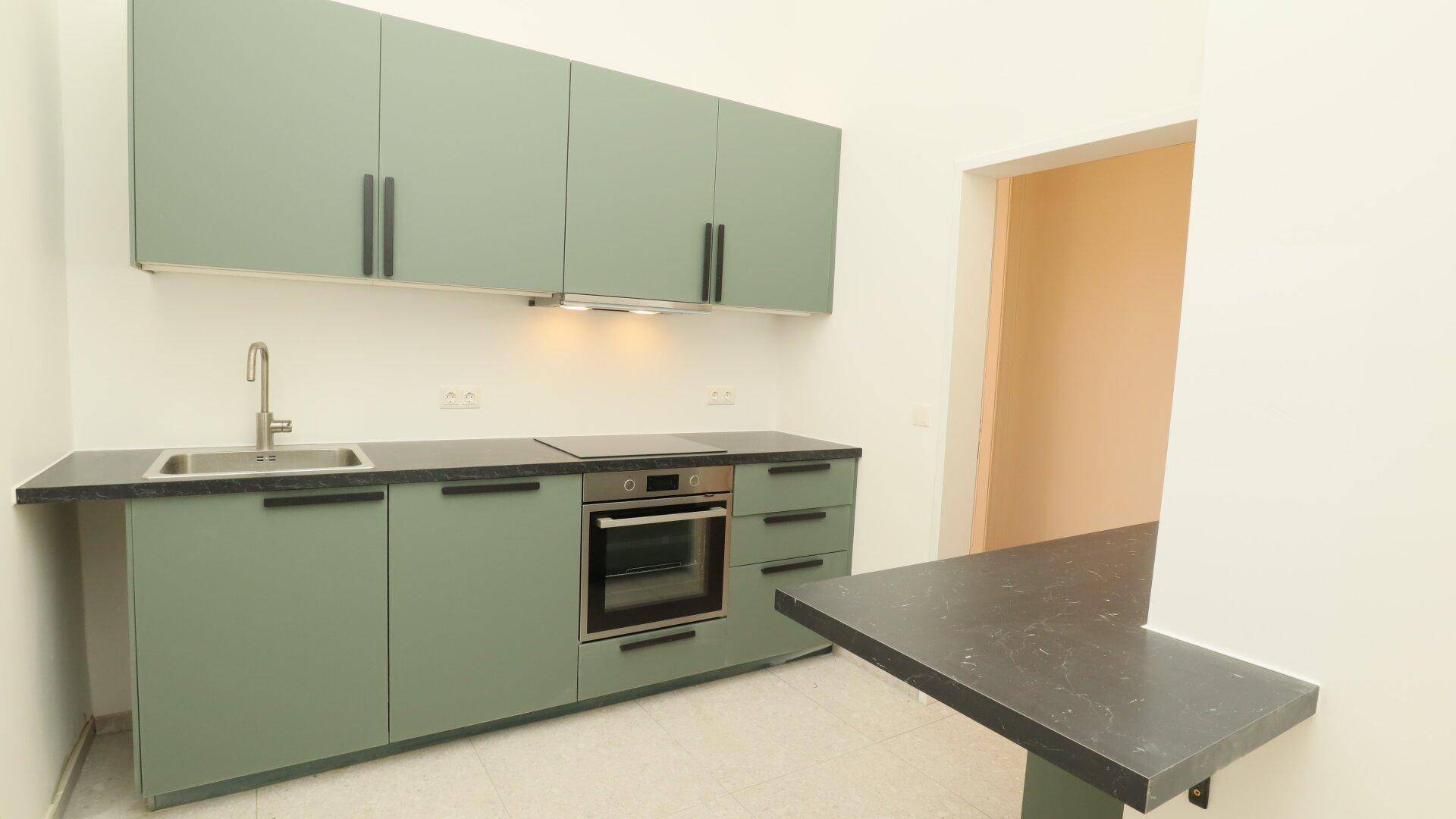 Moderne Küche mit grünen Schränken, dunklen Arbeitsplatten und integrierten Geräten, inklusive Kochfeld und Backofen.