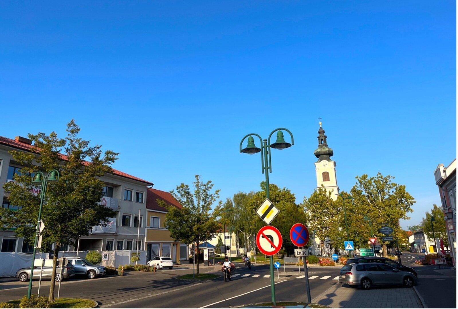 Blick auf eine Straße in Jennersdorf mit Gebäuden und einem Kirchturm im Hintergrund.