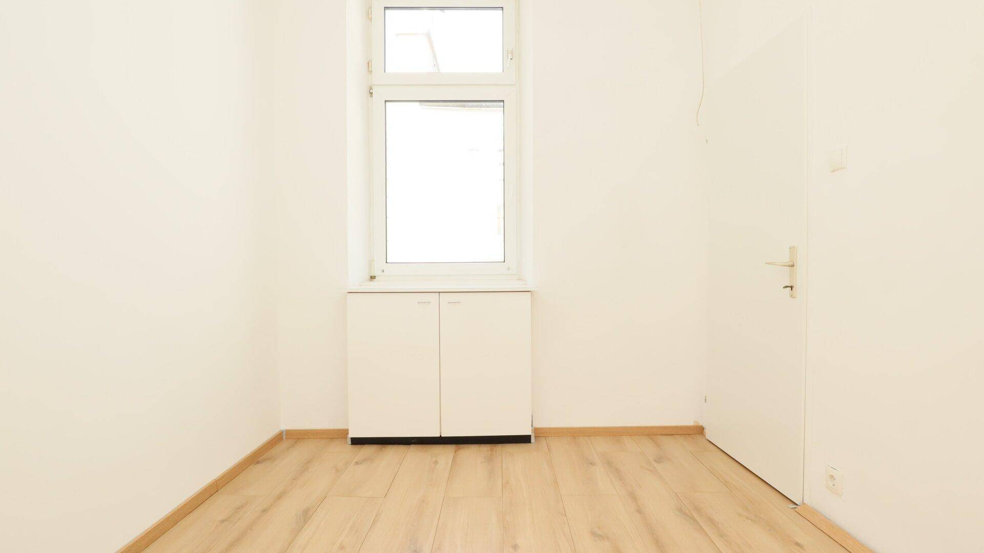 Kleiner, heller Raum mit einem Fenster, hellem Holzfußboden und einem weißen Schrank.